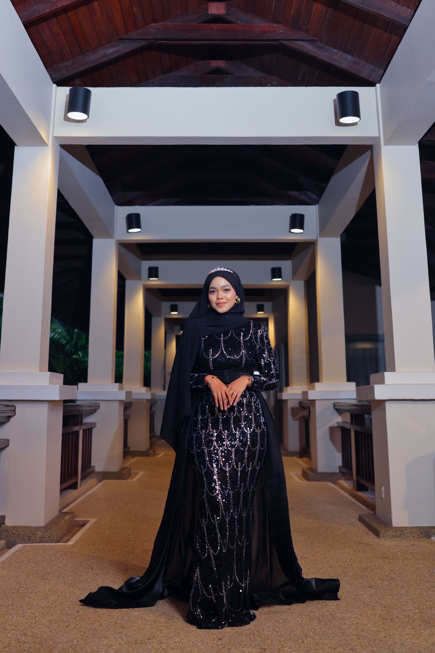 Elegant woman in black sequined hijab gown poses in illuminated hotel corridor at night.