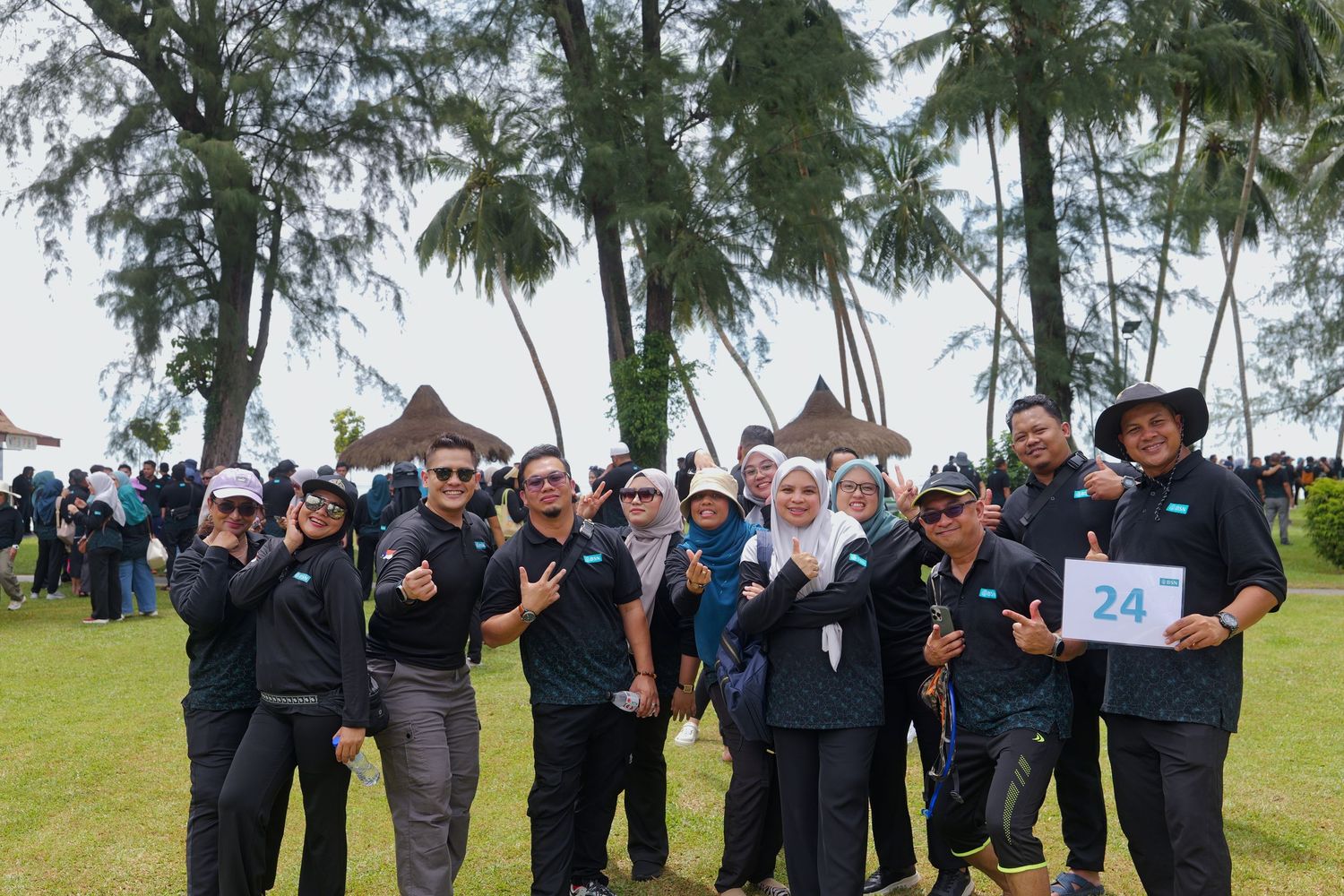 Group of smiling people in matching dark uniforms posing outdoors near tropical trees at a team event.