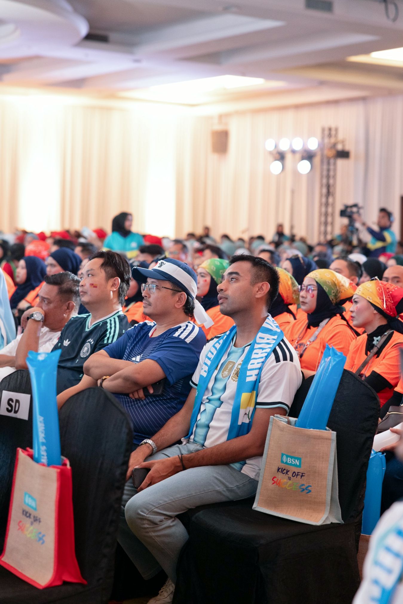 A diverse crowd of sports fans seated in a large hall, wearing colorful jerseys and holding branded tote bags at an event.