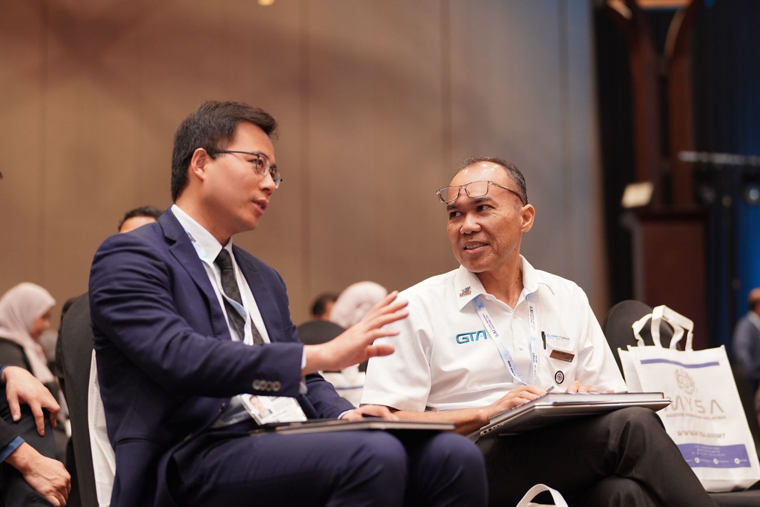Two attendees engage in conversation during a professional conference session while seated at a table.