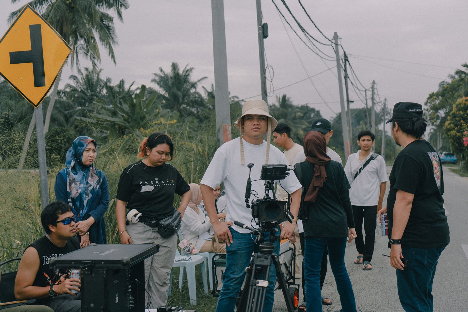 Film crew with camera equipment working on location outdoors alongside a palm tree-lined street.