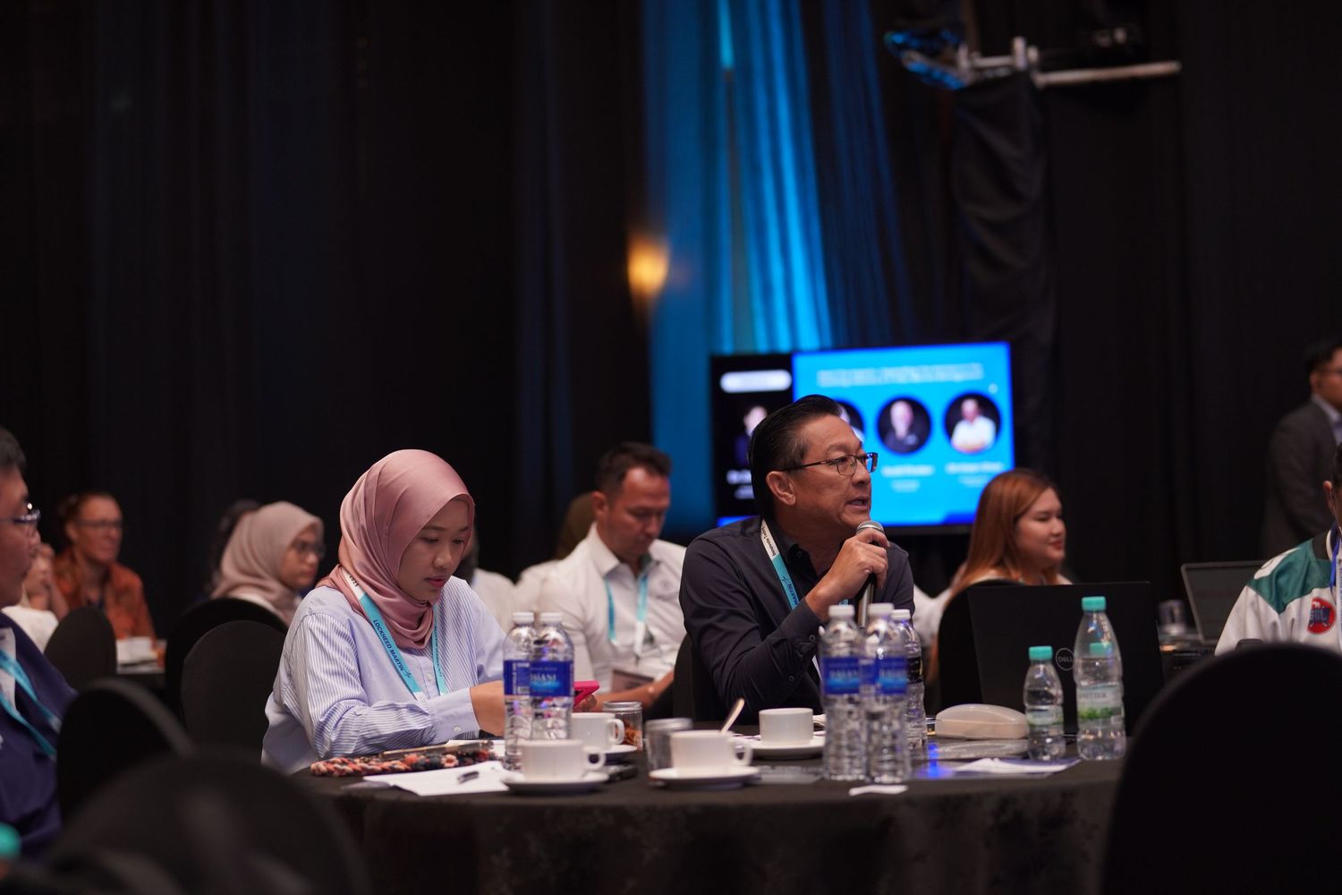 Diverse group of panelists seated at a long table during a conference presentation with blue backdrop lighting.