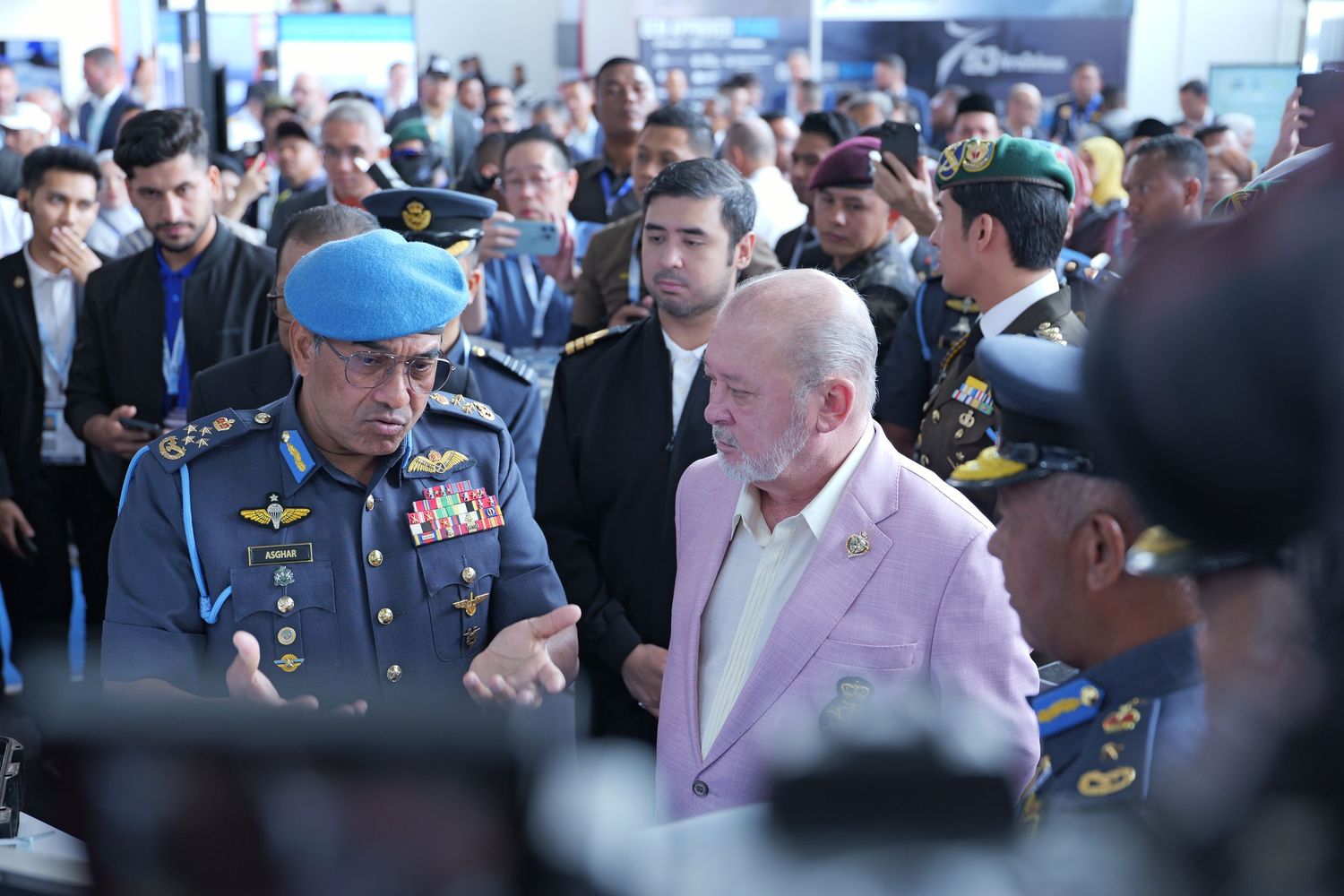A person in a pink suit jacket stands among a crowd with uniformed military personnel at an event.