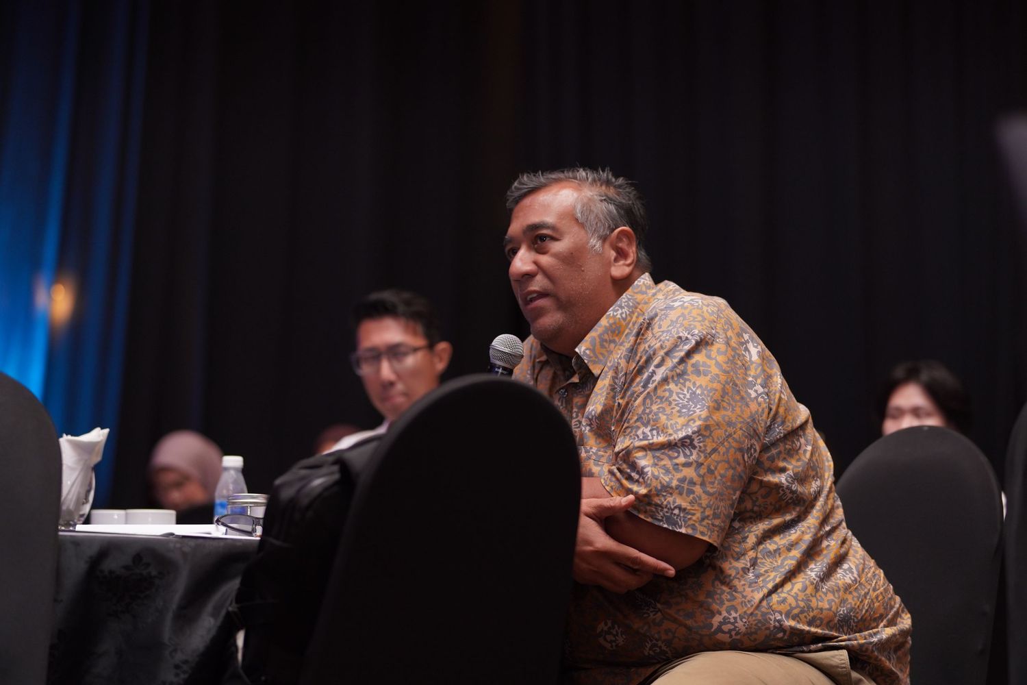 Speaker in a patterned shirt leans forward while speaking at a conference panel discussion.