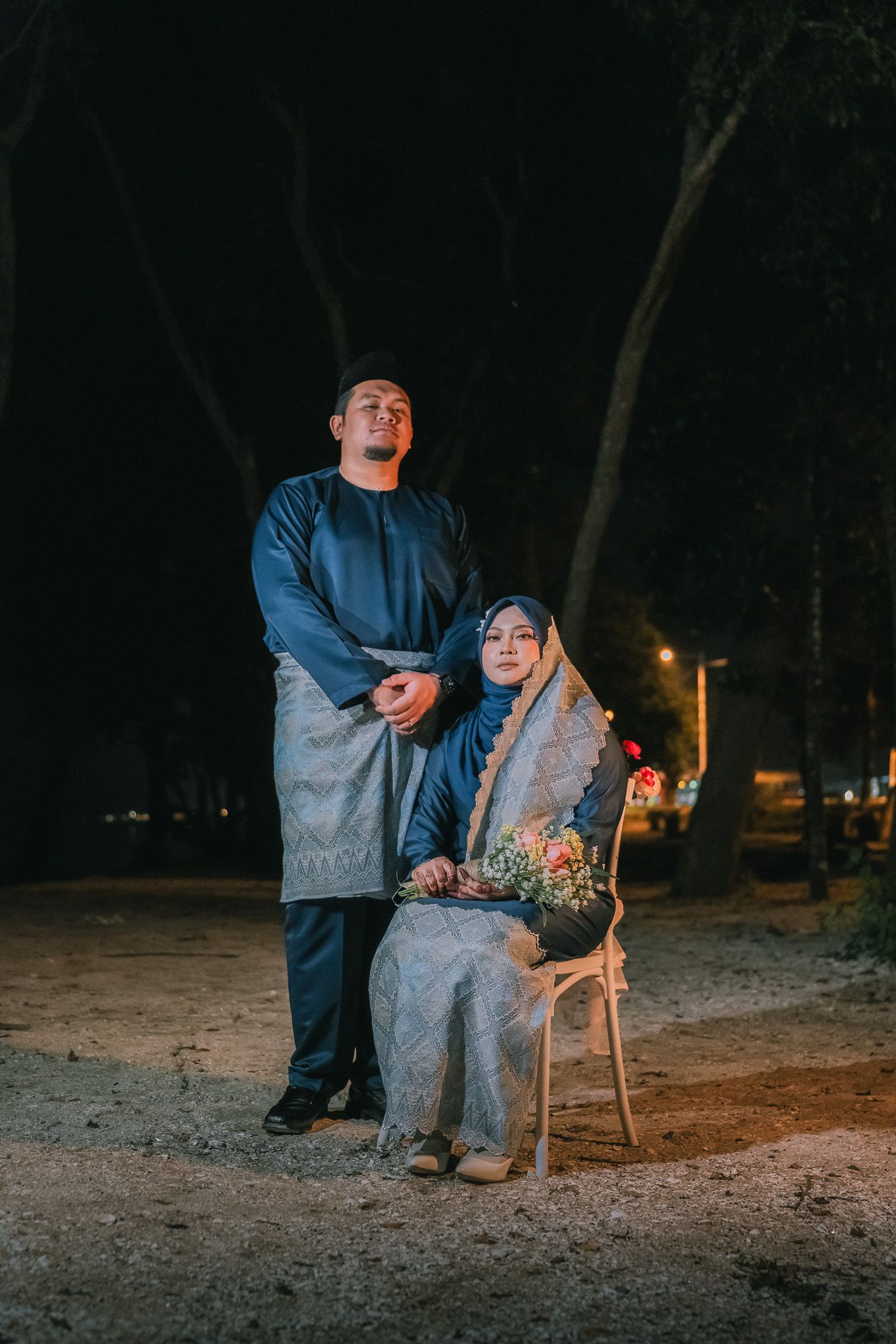 A couple in matching blue-grey outfits poses outdoors at night, with one person seated holding flowers in a moody setting.