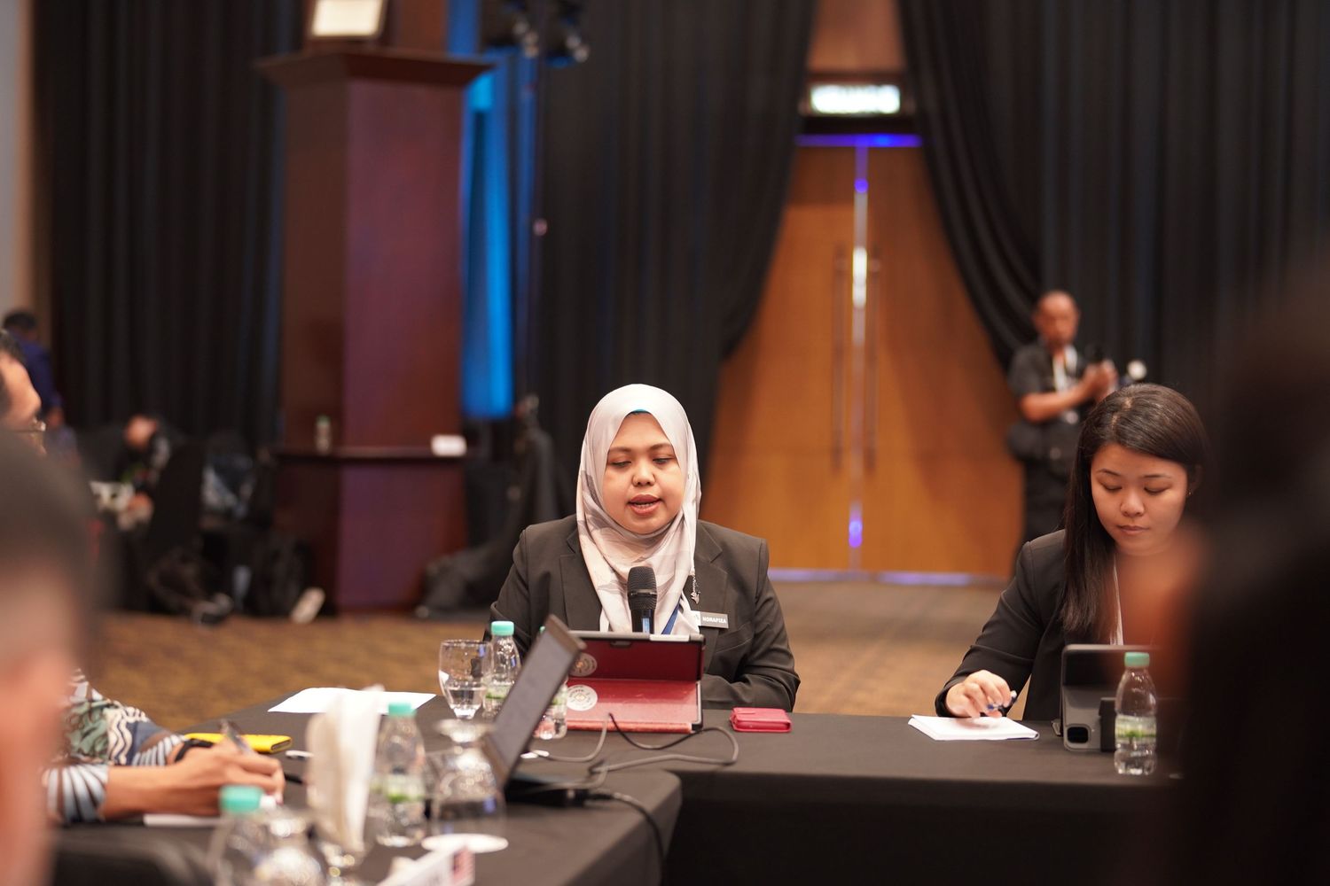 Conference attendees participate in a roundtable discussion in a formal meeting room setting.