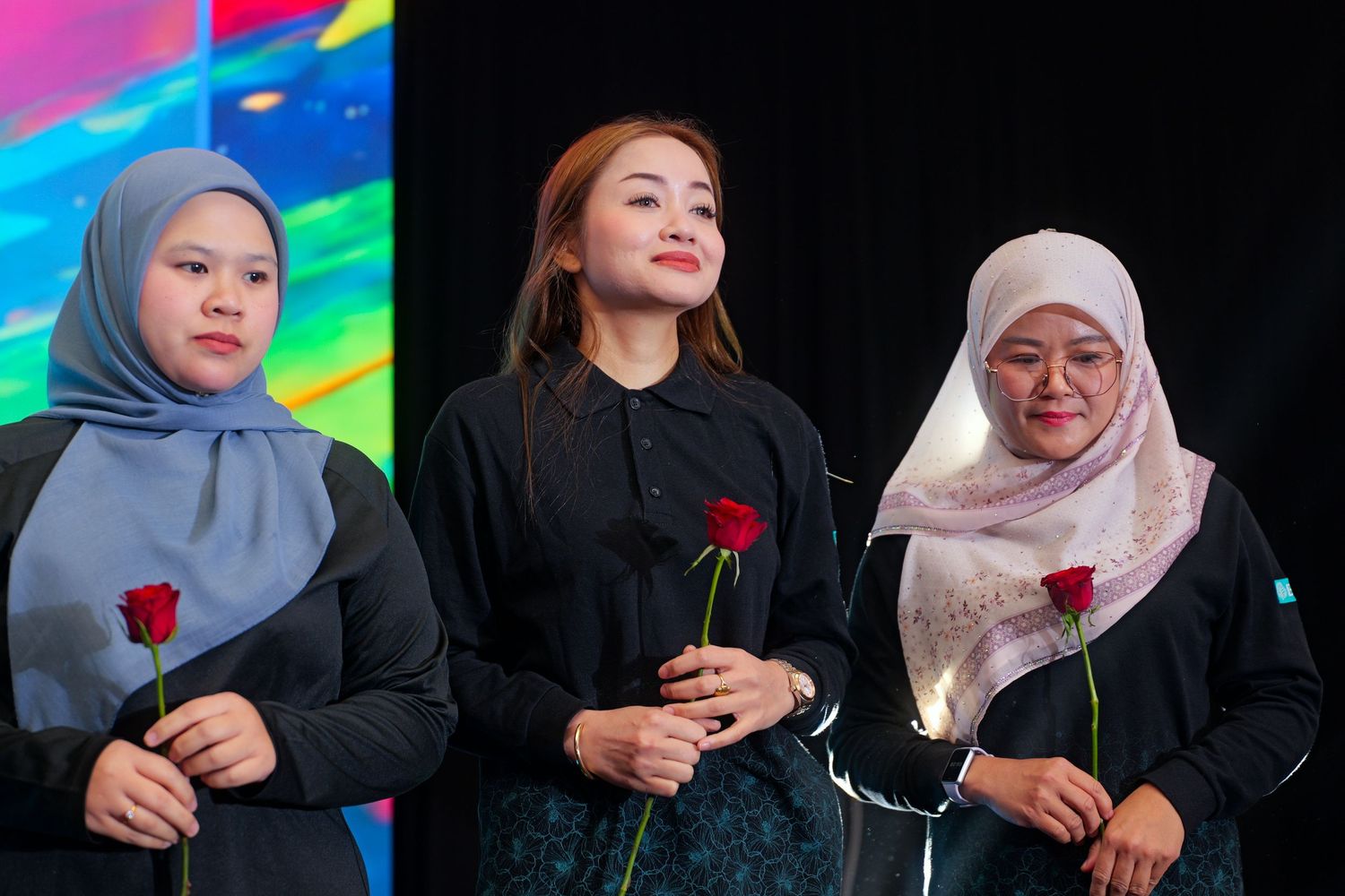 Three women holding red roses standing on stage in front of a colorful backdrop.