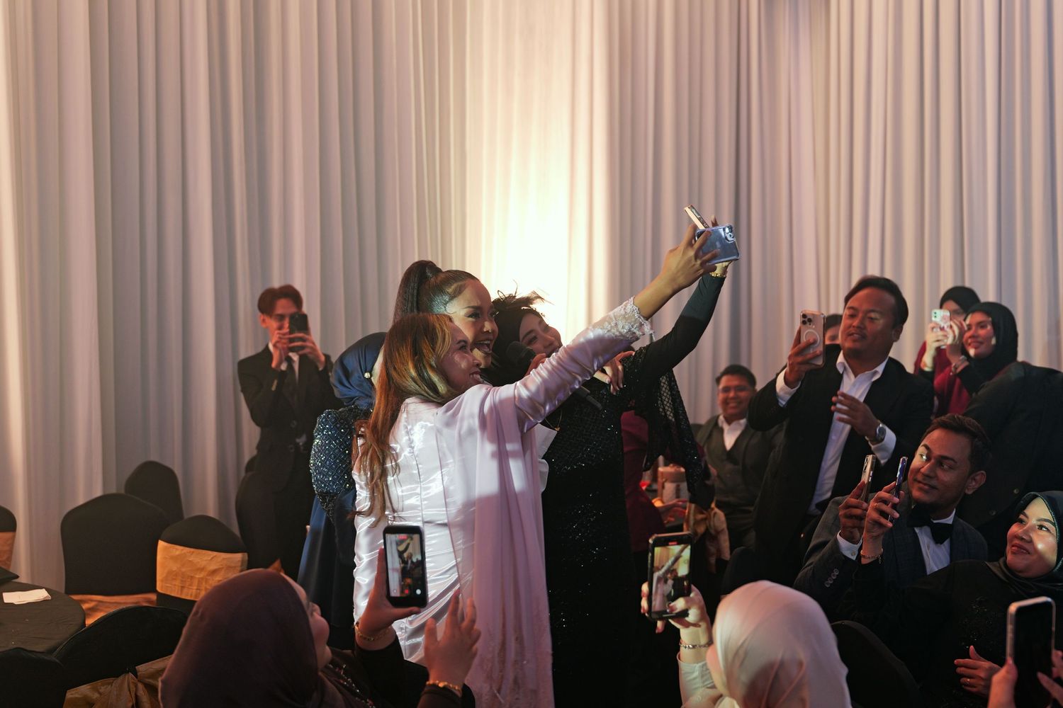 Crowd taking selfies with a performer at a lively indoor celebration or awards ceremony event.