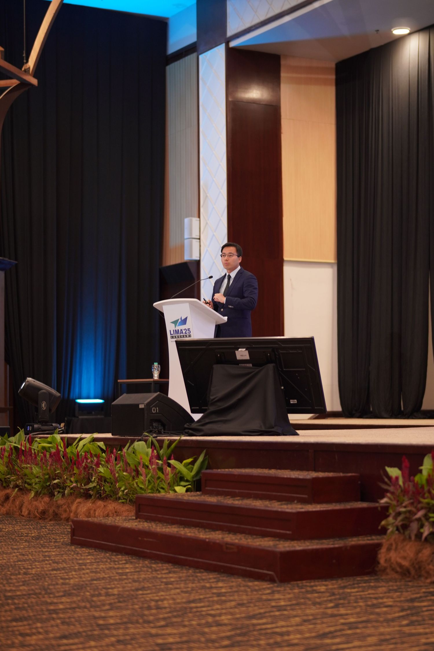 A speaker stands at a podium on an elevated stage with dark curtains and decorative plants.