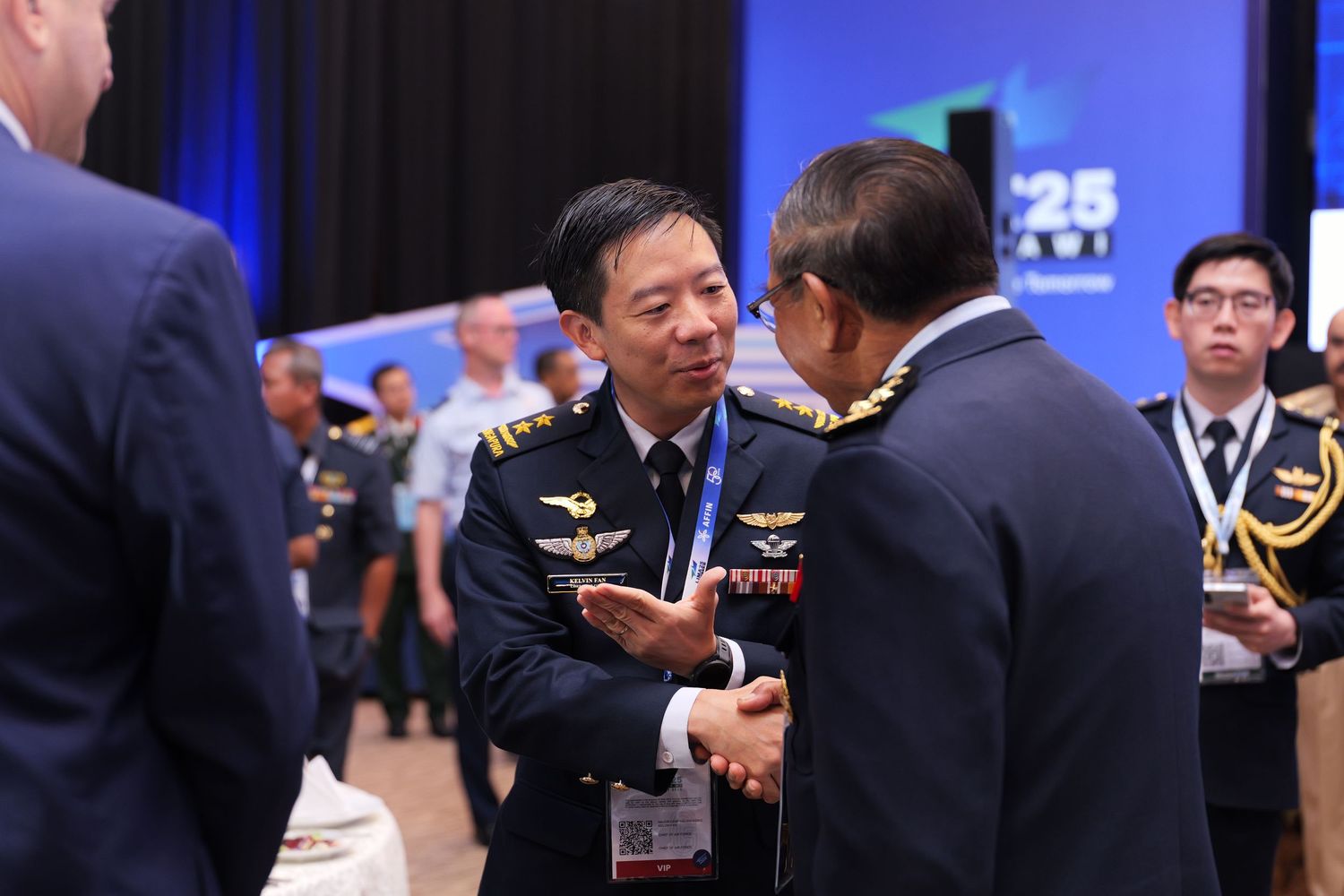 Military officials exchange greetings at the LIMA aerospace conference with blue backdrop.