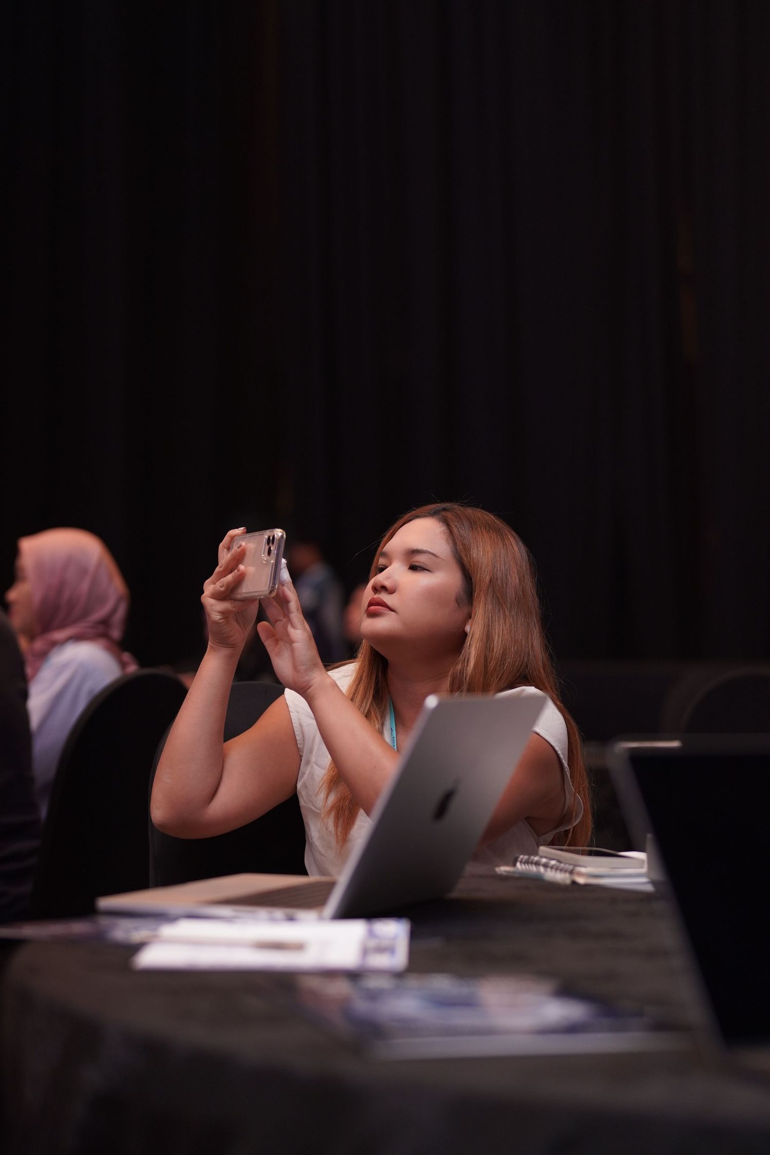 Woman in white top using phone while seated at conference table with laptop.