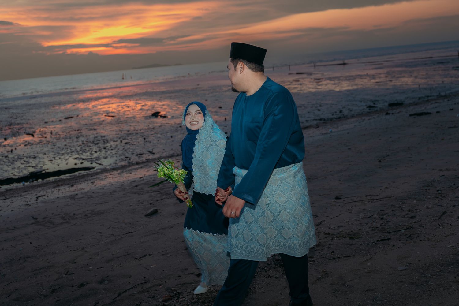 Two people walk along a beach at sunset with colorful orange and pink clouds reflecting on the ocean water.