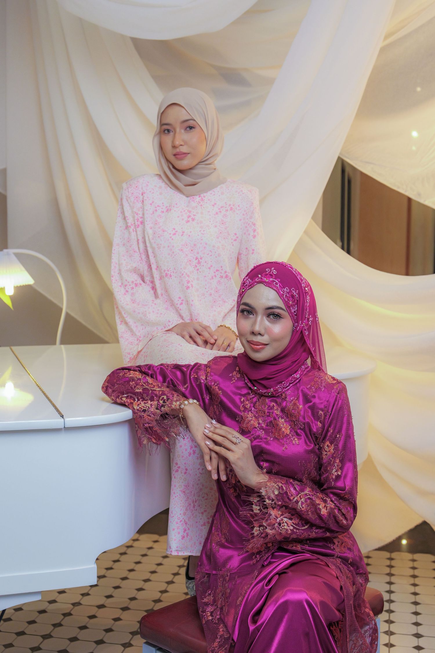A woman in a vibrant magenta hijab and formal dress poses elegantly against a white draped backdrop.