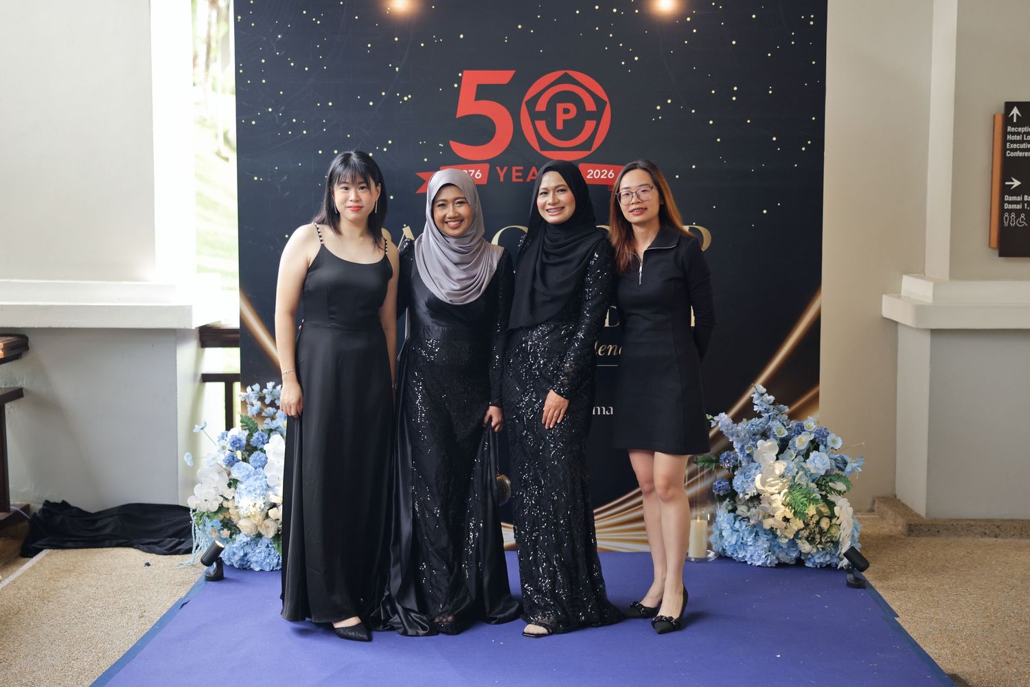 Four women in elegant black attire pose together at a 50th anniversary celebration event with floral decor.