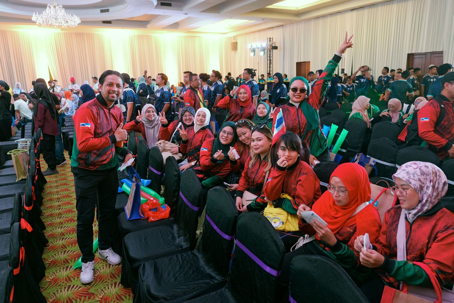 Large group of people in red and green outfits cheering enthusiastically at an indoor event.