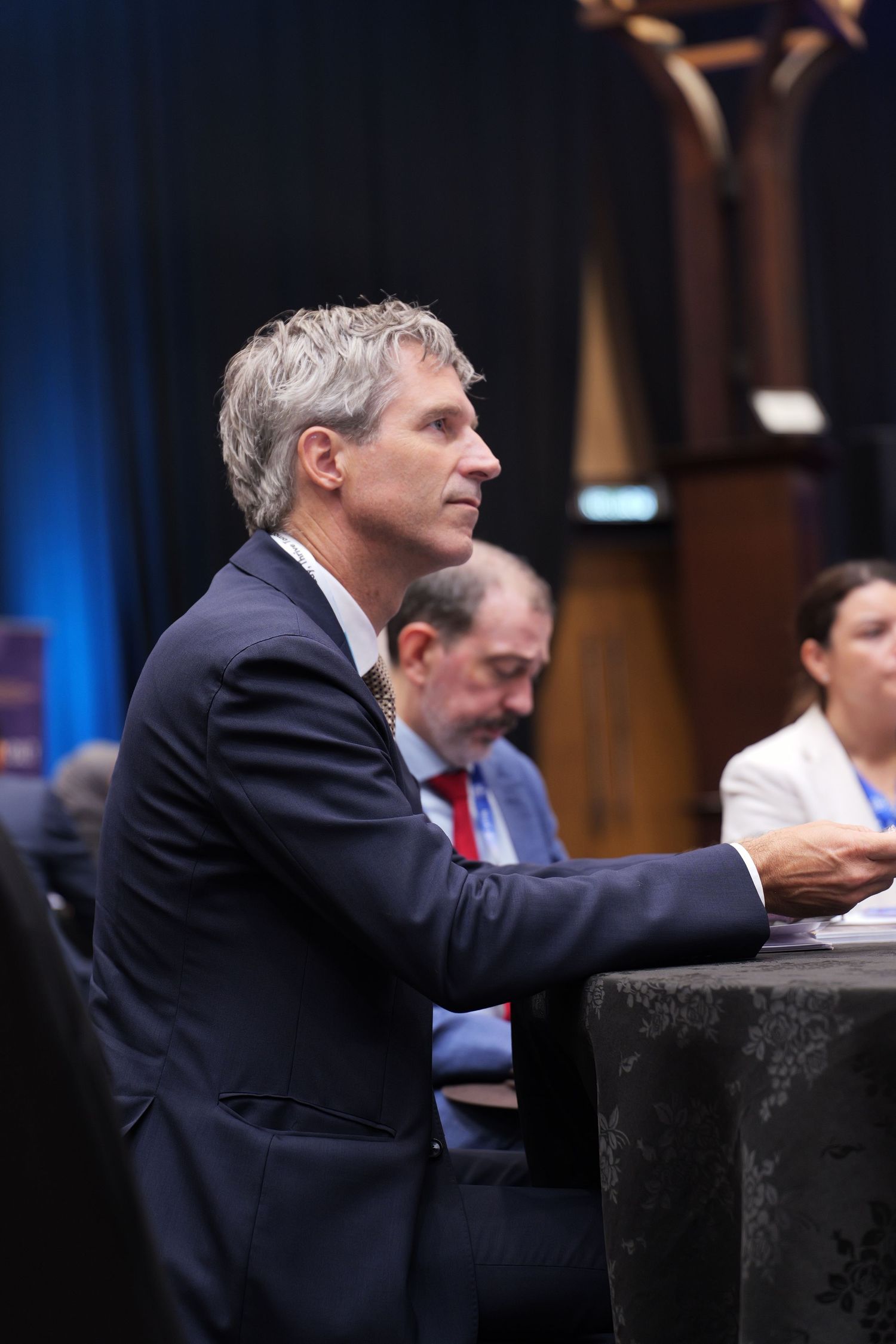 Professional in a dark suit gestures while speaking at a conference podium against blue background lighting.