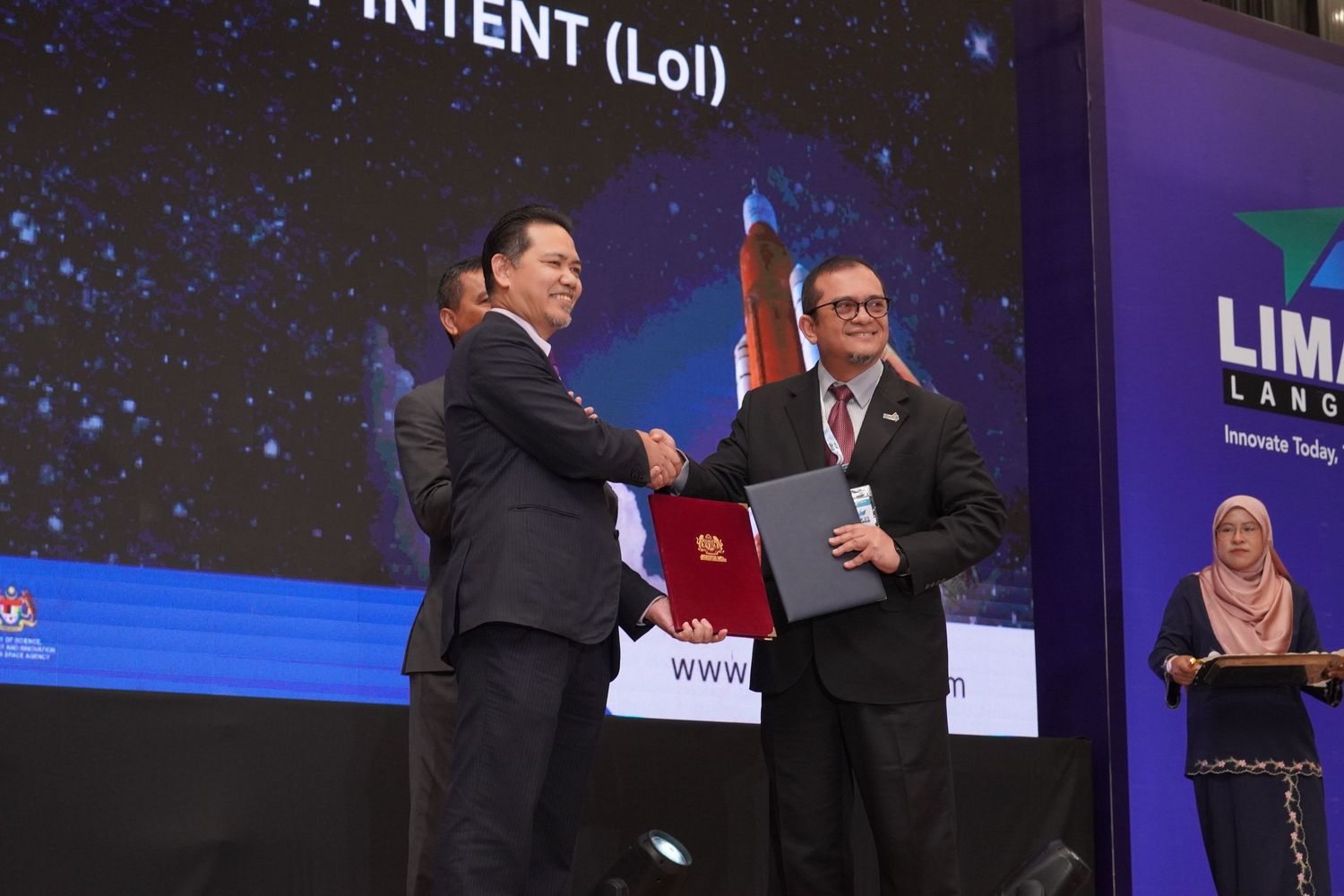 Two people shaking hands during an award ceremony presentation on stage with a large display screen in the background.