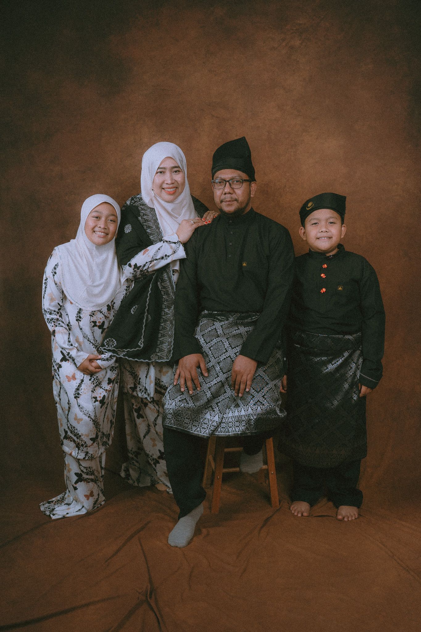 A family wearing traditional Malay formal attire poses together against a brown studio backdrop.