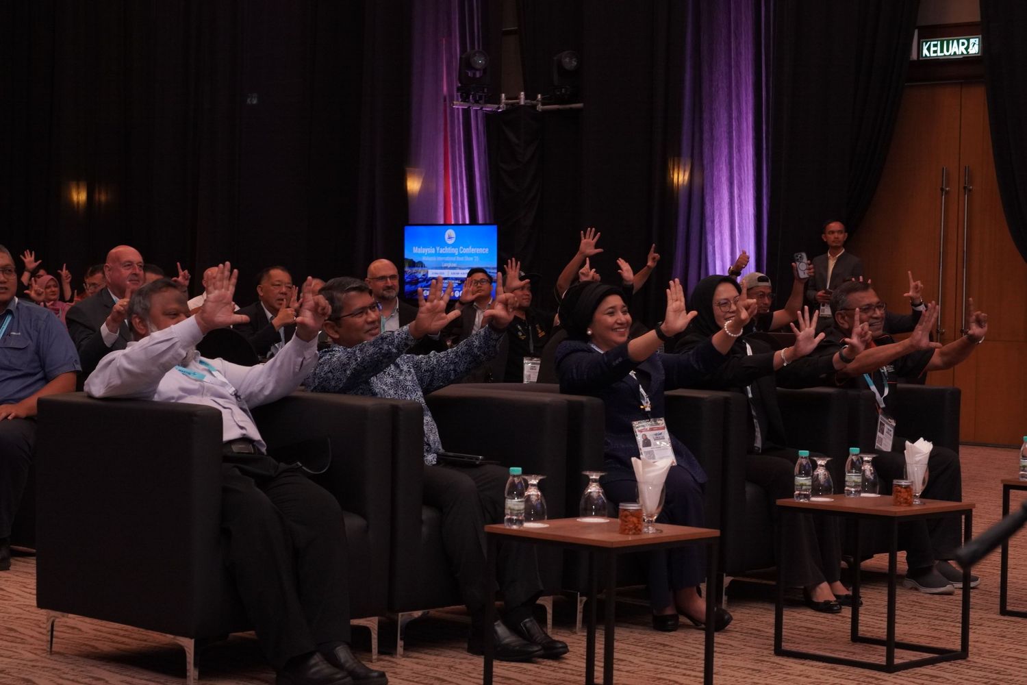 People sitting in rows on a stage raise their hands during what appears to be a conference or panel event.
