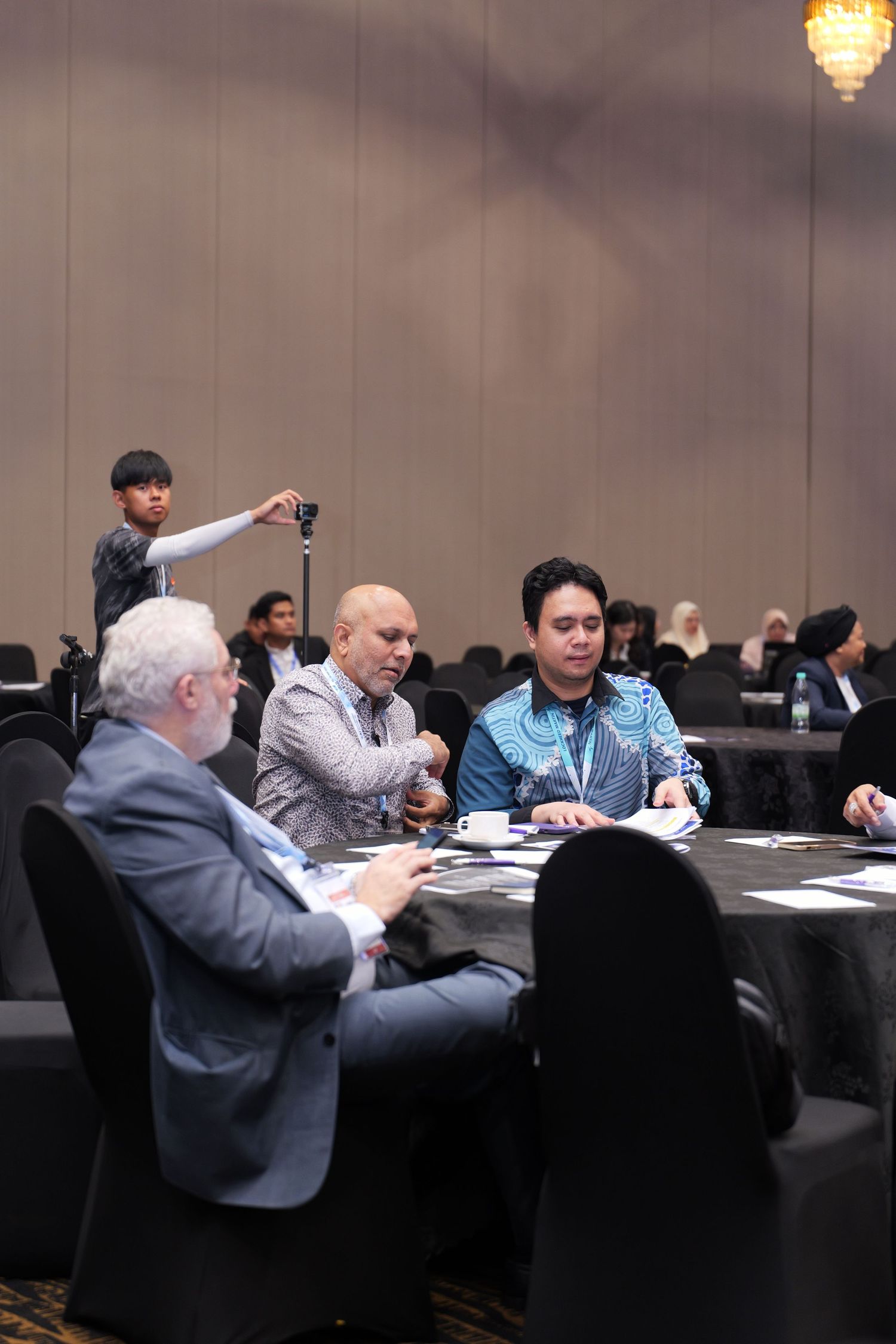 People seated at a round table during a conference or business meeting in a hotel conference room.