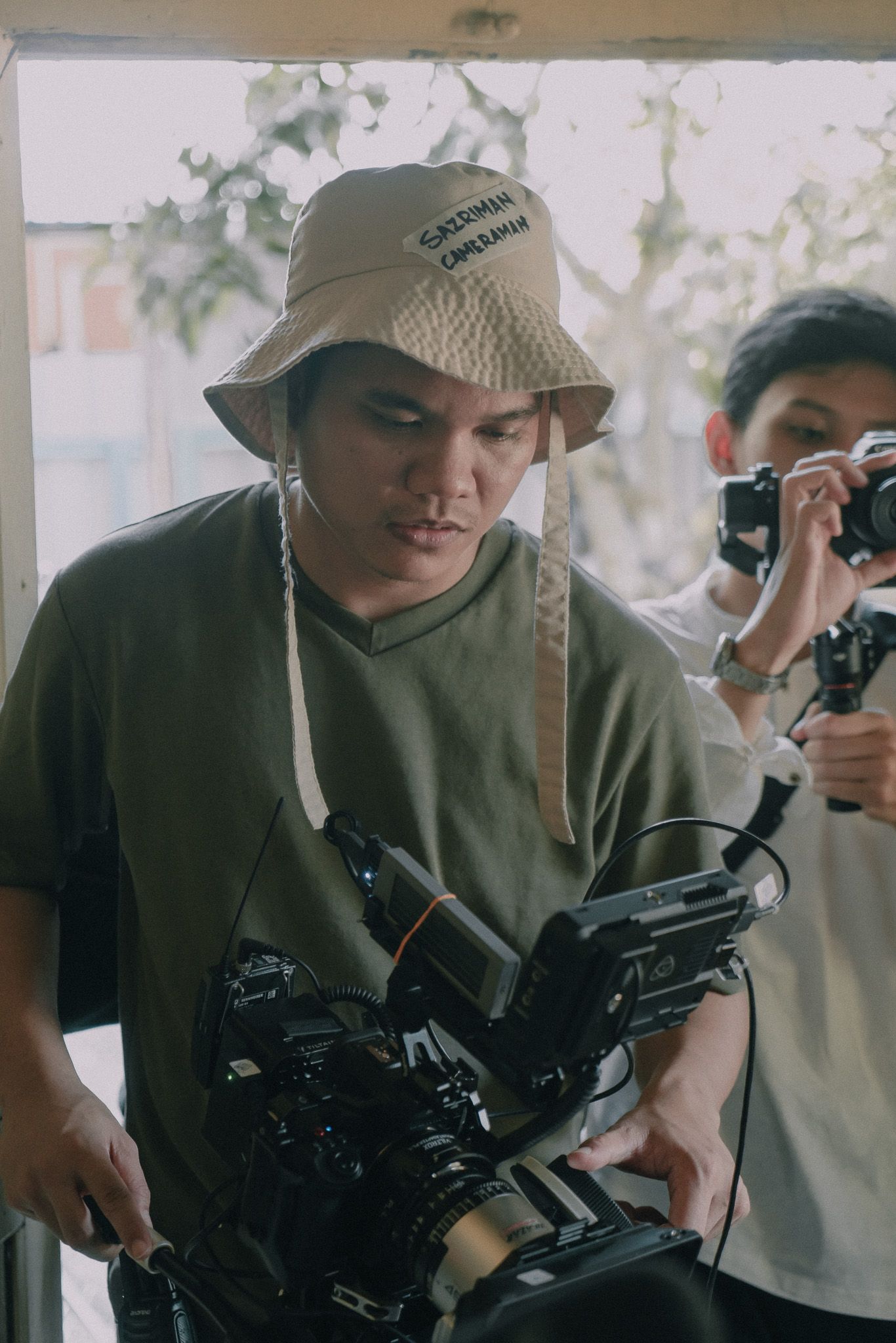 Camera operator wearing a bucket hat and green sweatshirt holds professional video equipment while filming.