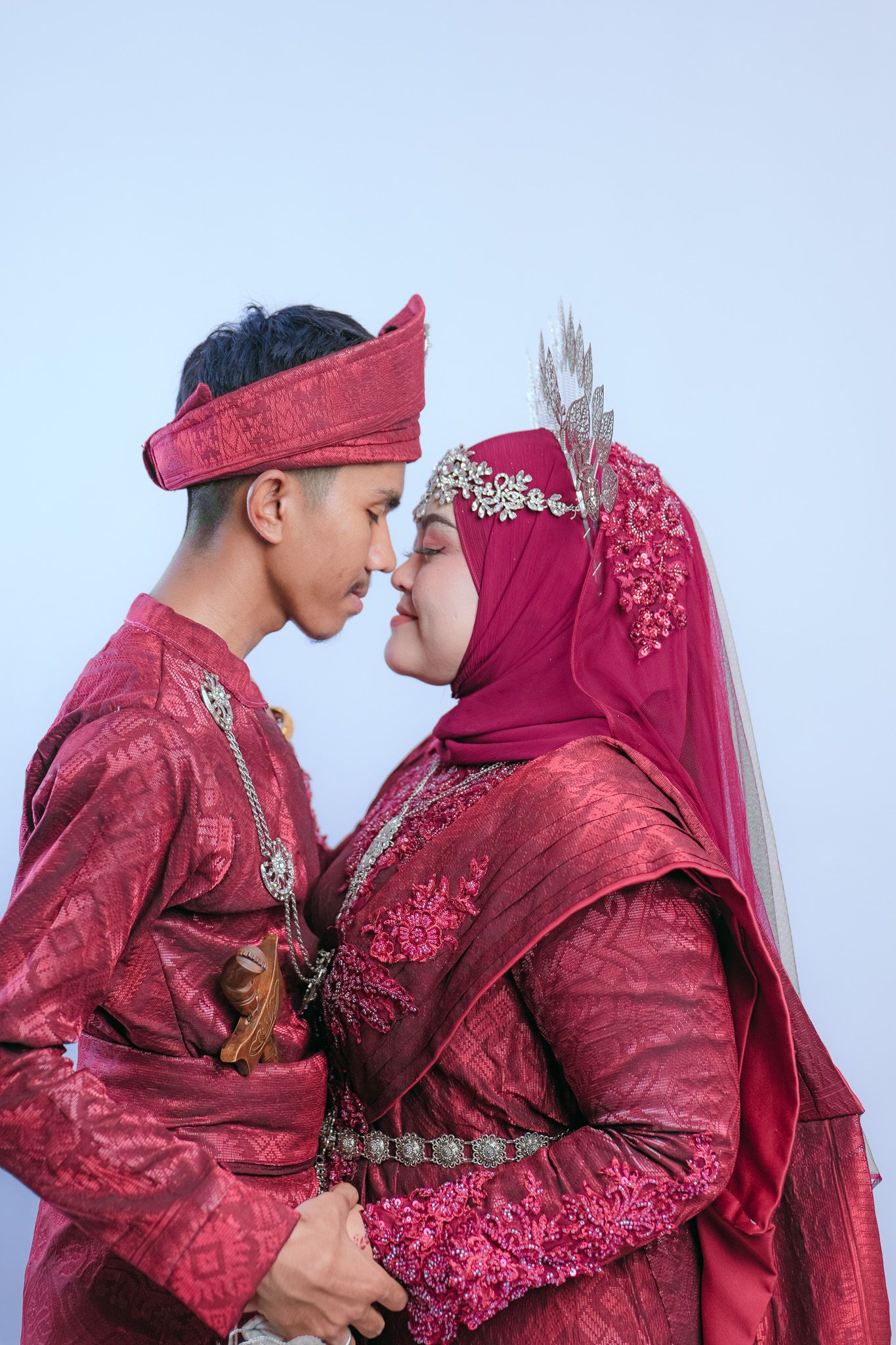 A couple in matching deep red traditional Malay wedding attire pose together against a light background.