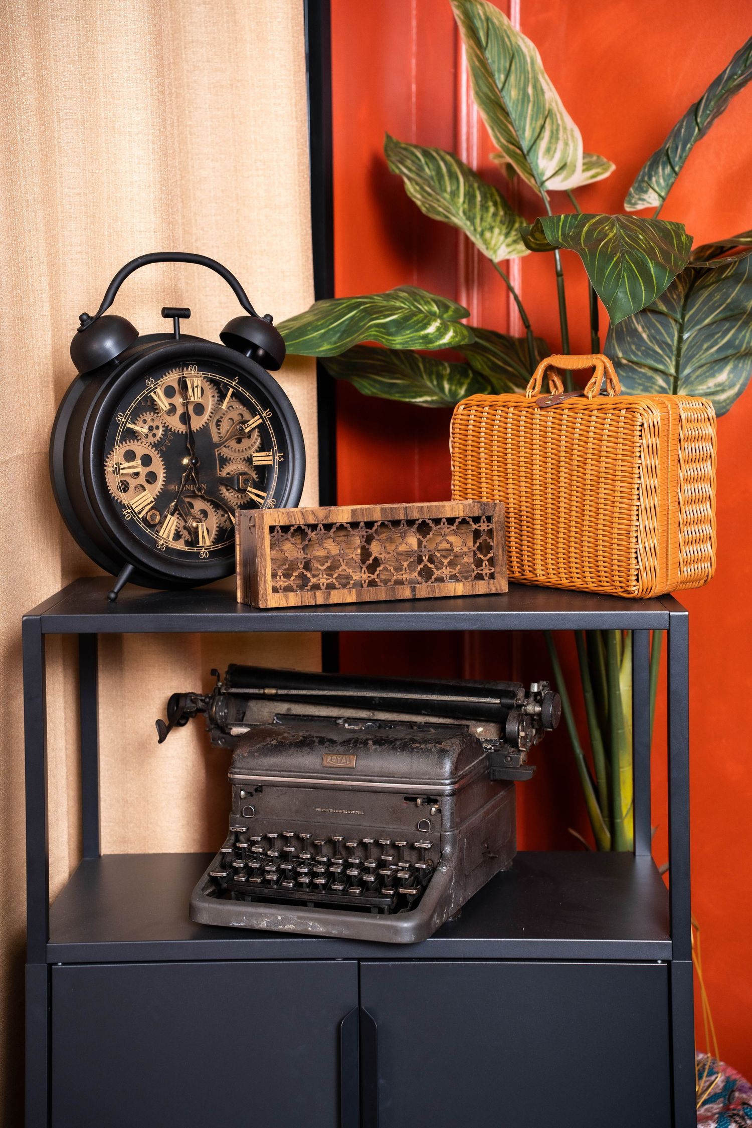 Vintage black typewriter displayed on dark cabinet with orange wall background and decorative plant accents.