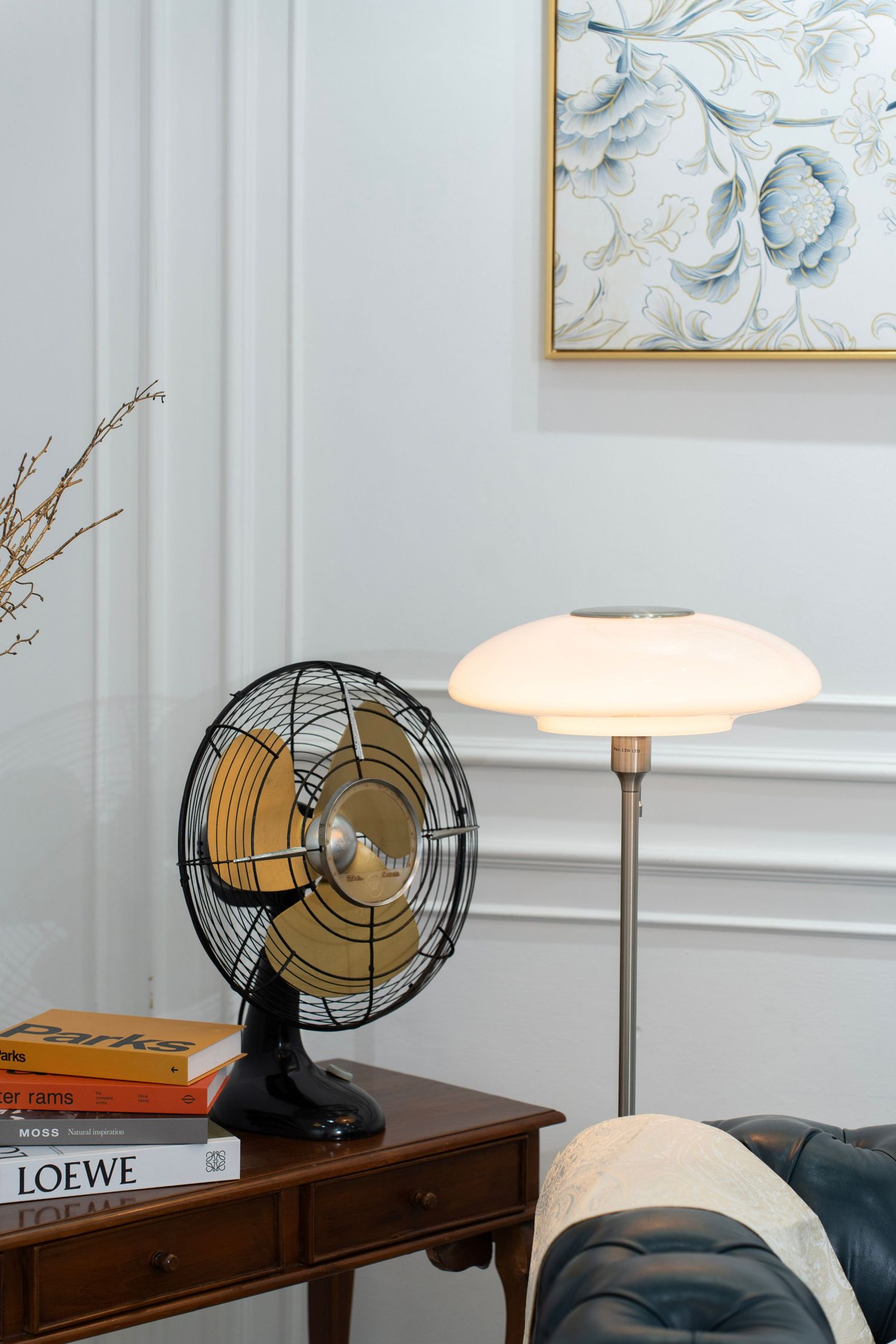 Vintage black metal desk fan next to a modern table lamp with white shade on wooden furniture against light walls.