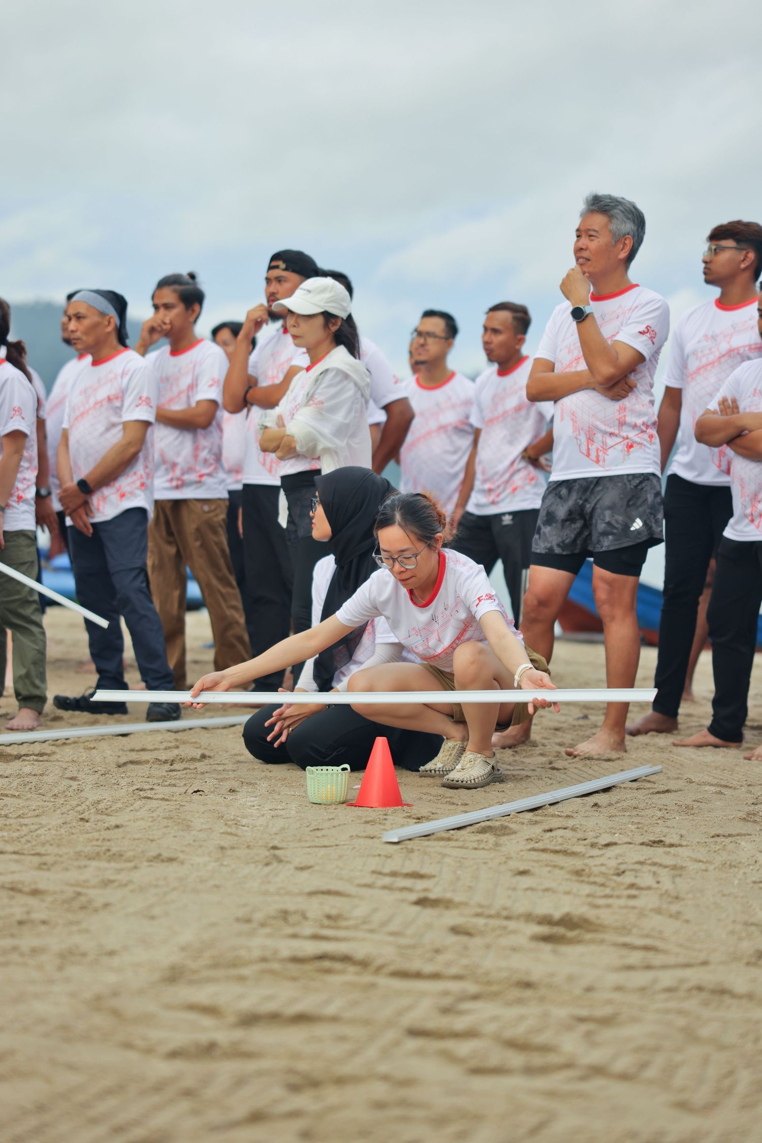 People in white shirts participate in a beach team building activity measuring sand with poles and orange cones.