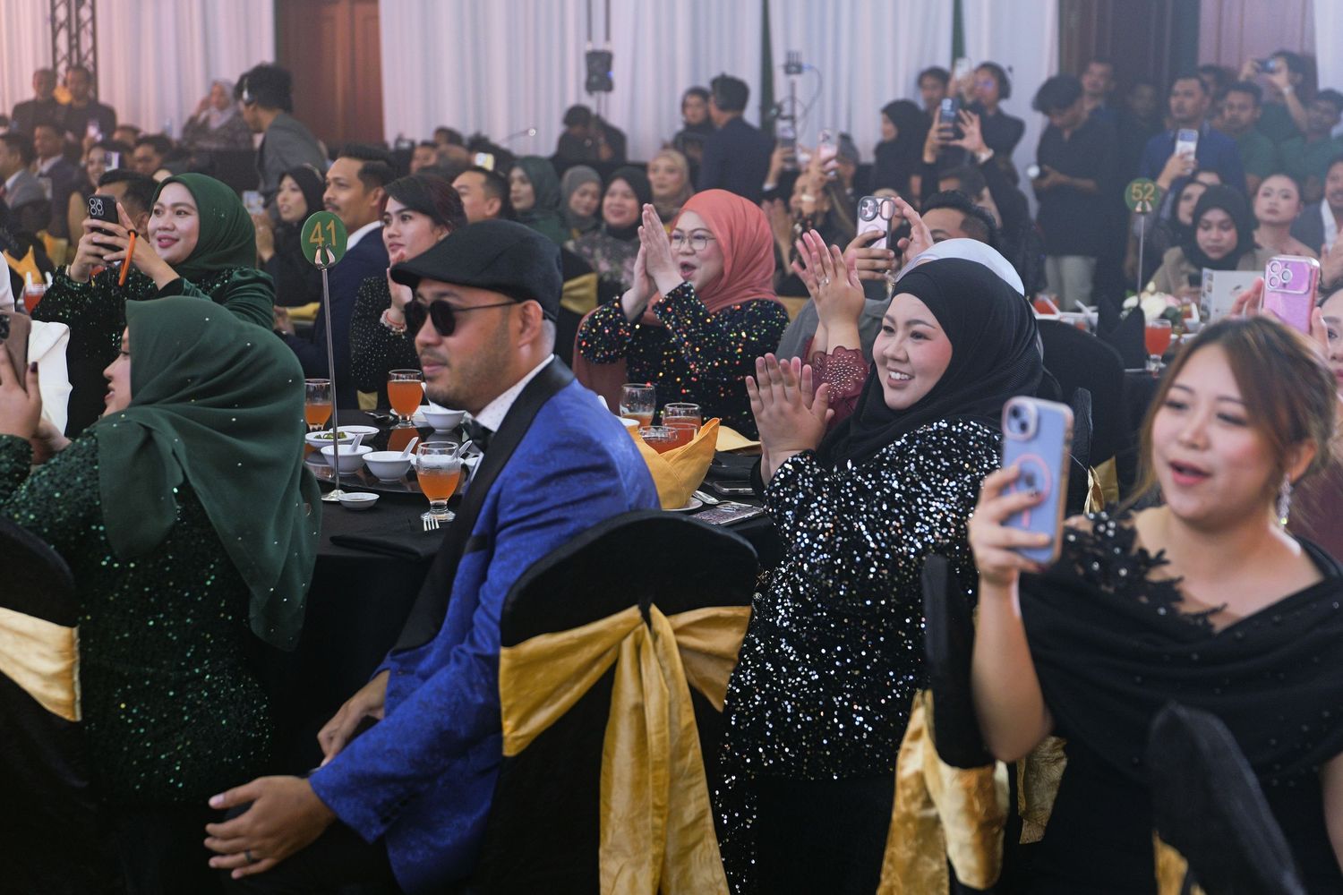 Guests in black formal attire applauding and taking photos at a lively corporate gala dinner.