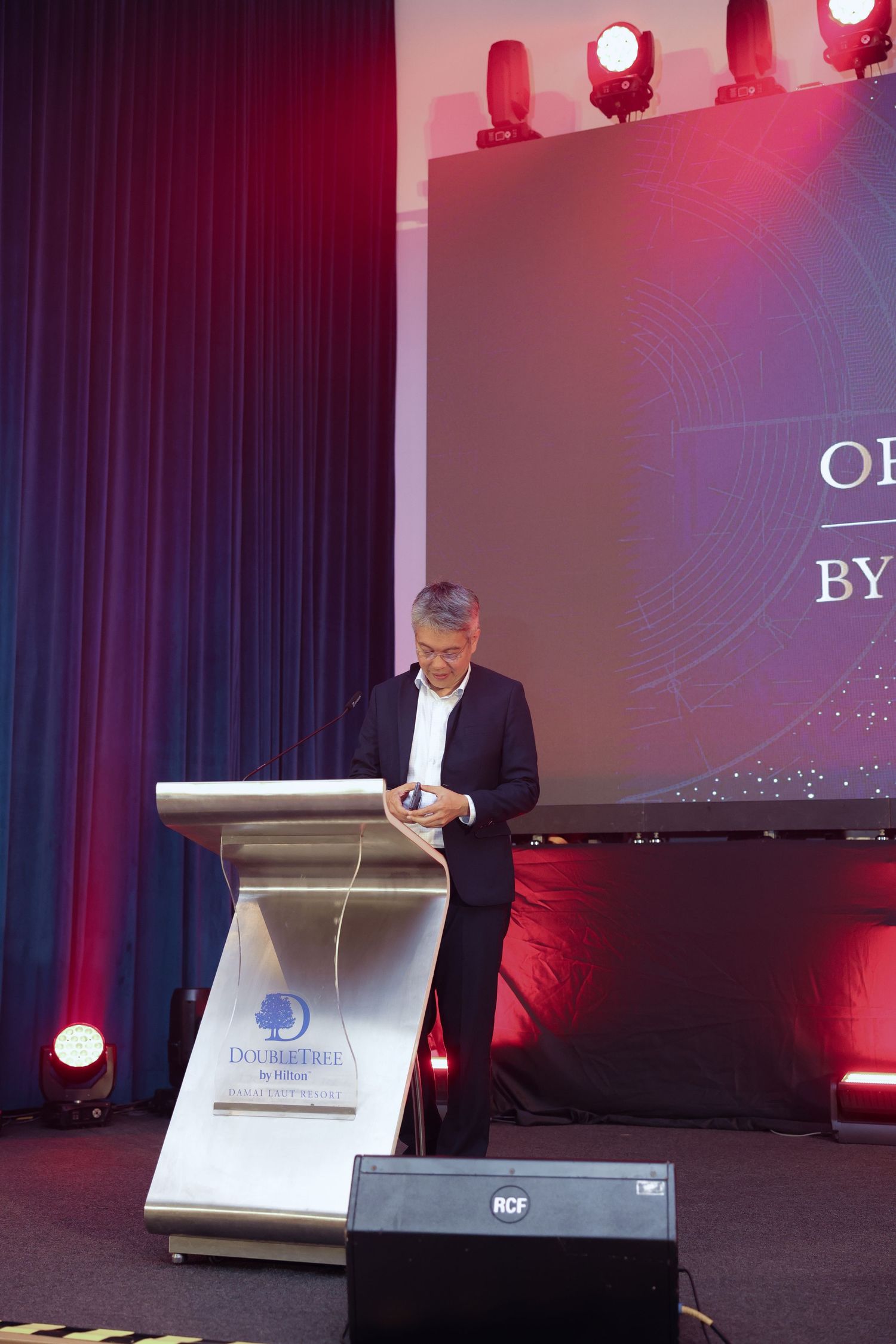 A man in a suit speaks at a podium at a DoubleTree hotel corporate event with stage lighting.