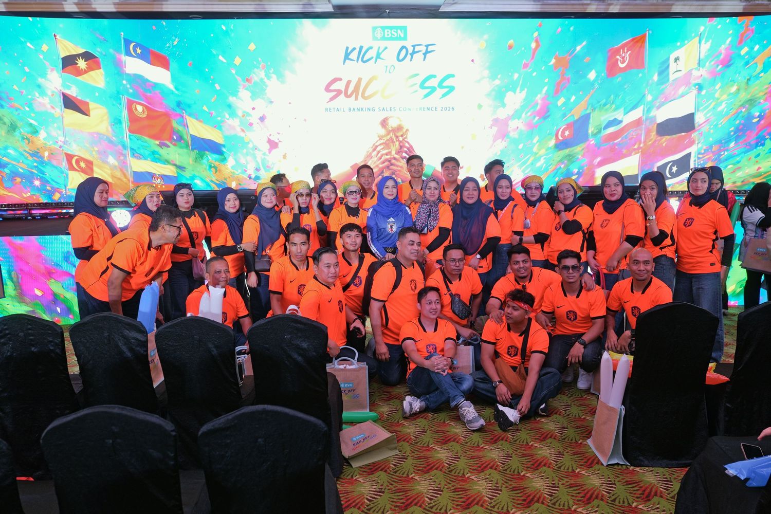 Large group in orange uniforms poses in front of a colorful Kick Off Success event backdrop.