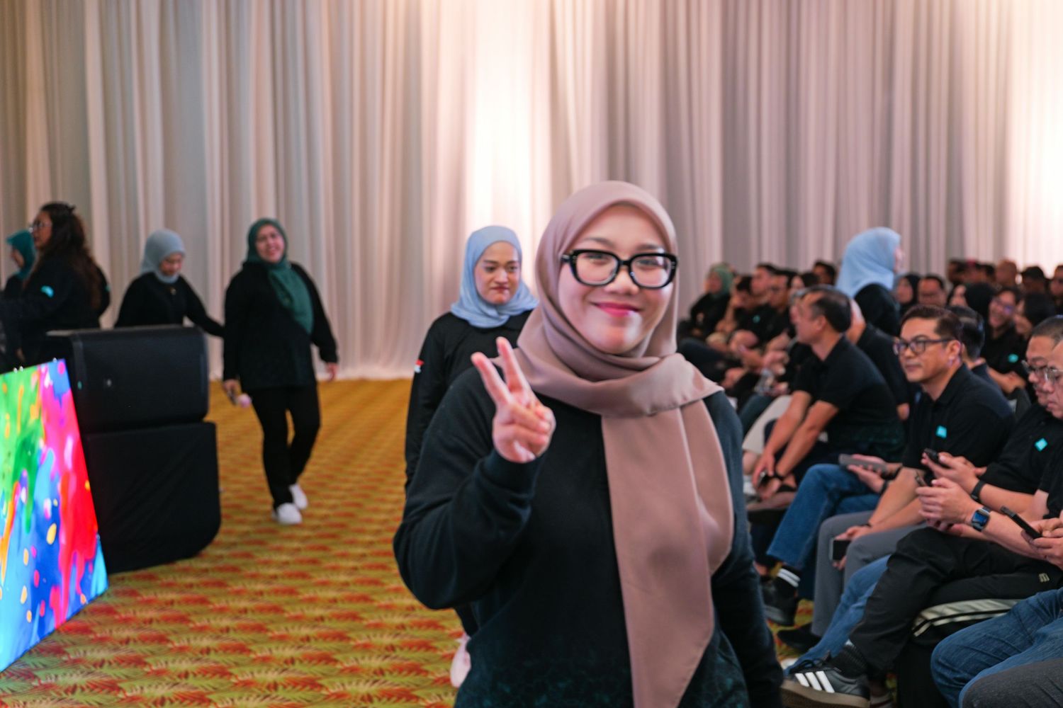 Smiling woman in hijab and glasses making a peace sign gesture at an indoor event.