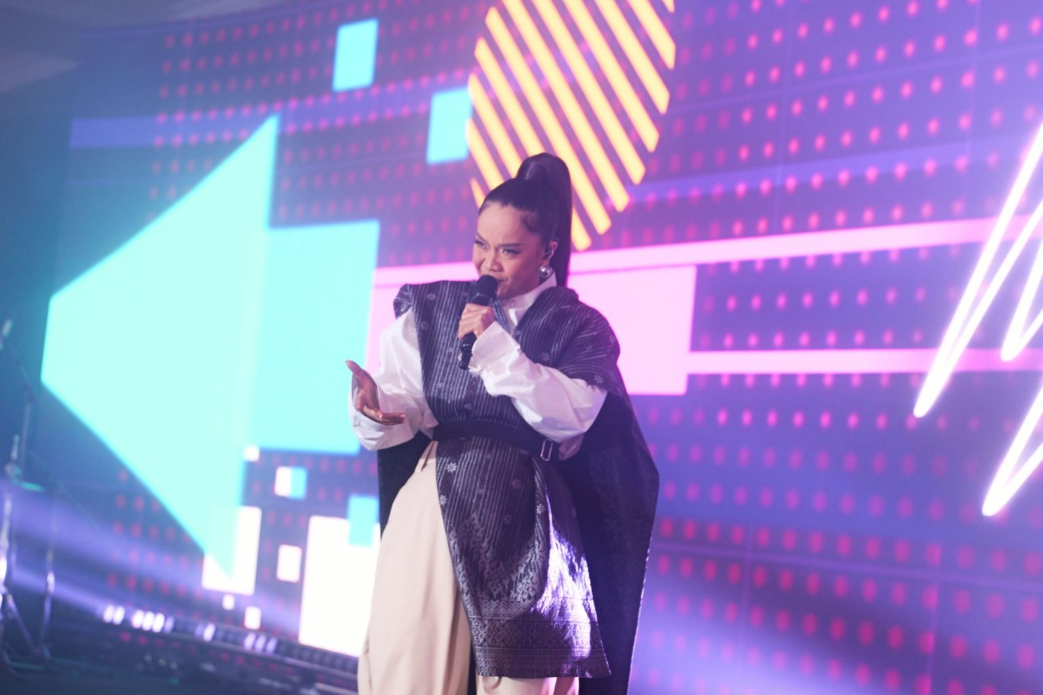 Performer in stylish black and white outfit speaking into microphone on a colorful lit stage.