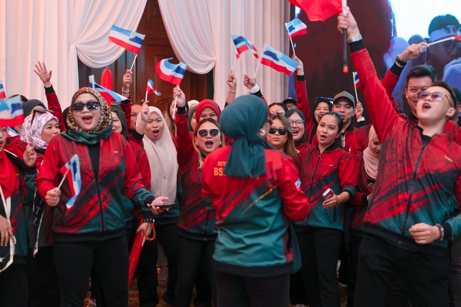 Enthusiastic group in red uniforms waving Malaysian flags celebrating at an indoor event.