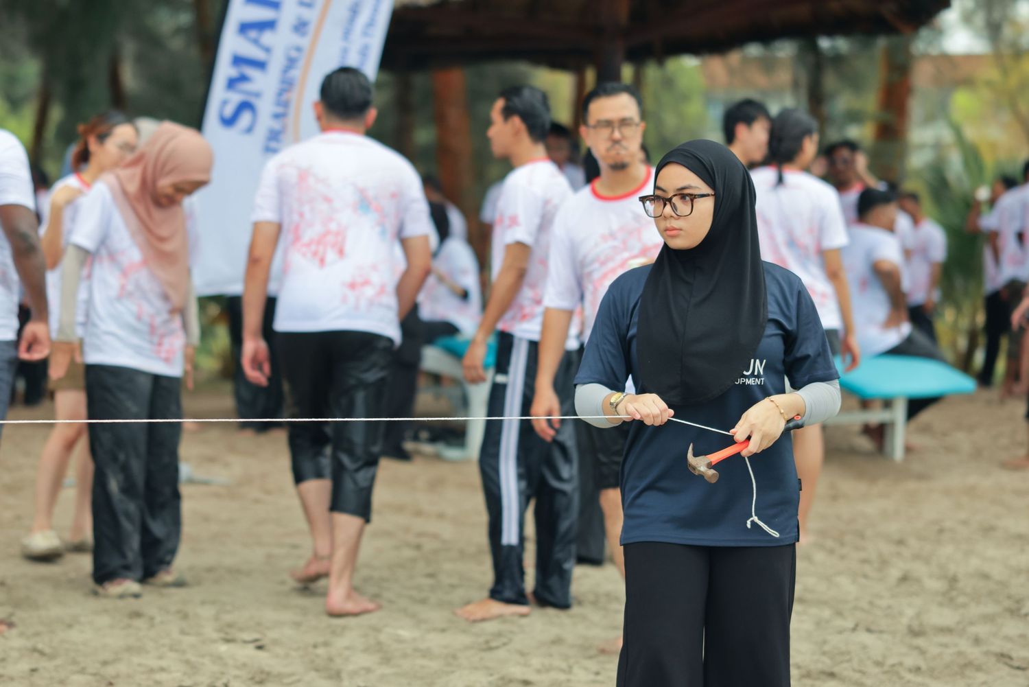 Woman in hijab holding string during outdoor team building activity on sandy ground with group in white shirts.
