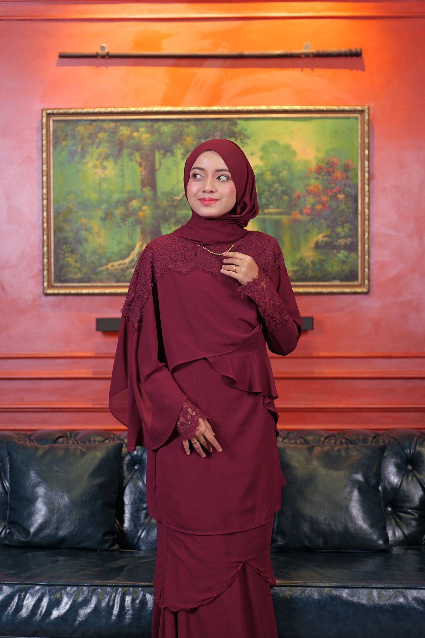 A person wearing a burgundy hijab and matching layered dress stands in front of a framed artwork on a red wall.