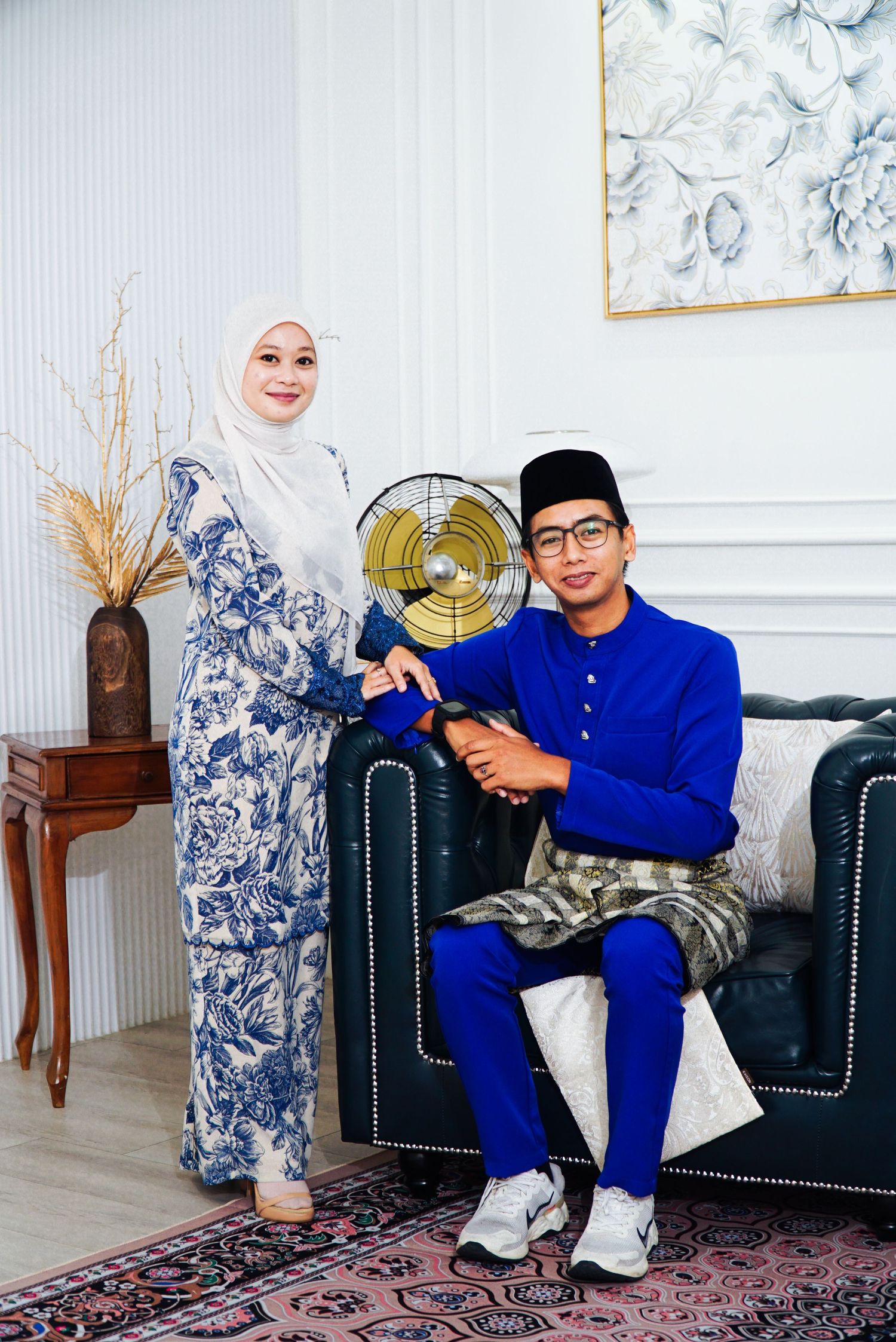 A couple in traditional Malay attire sits in an elegant living room with blue and white decor and oriental rug.
