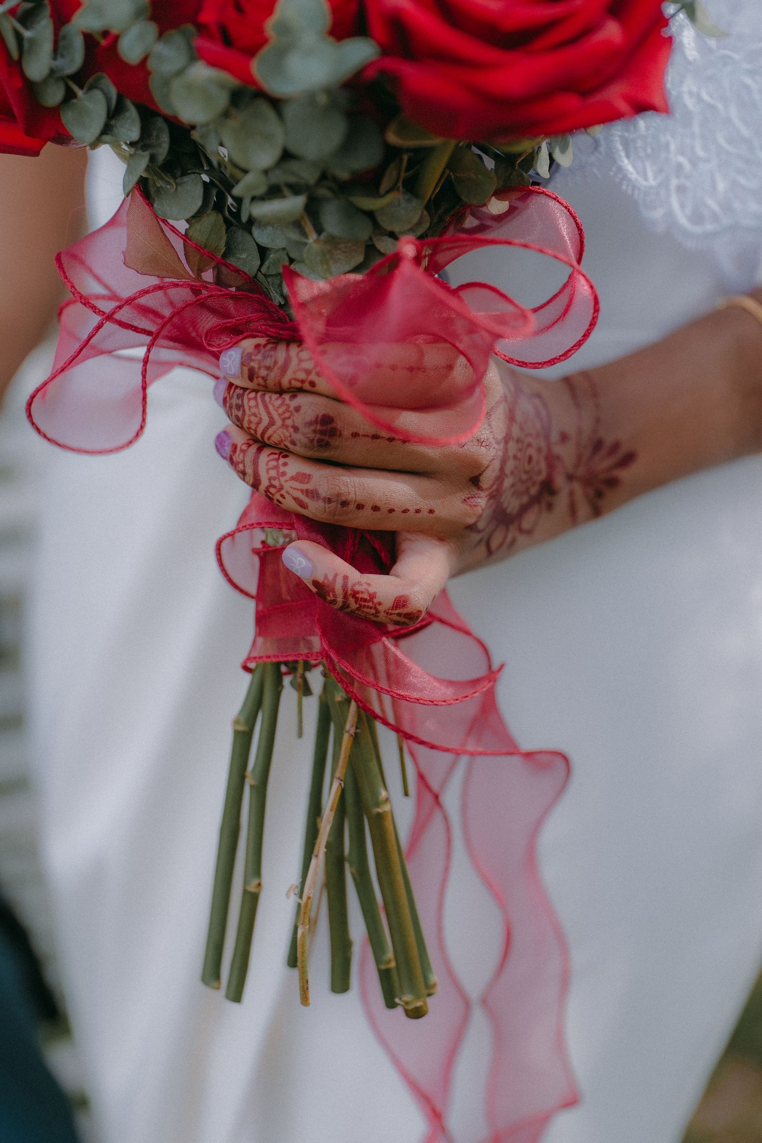 A bridal bouquet of red roses tied with ribbon is held by hands decorated with henna.