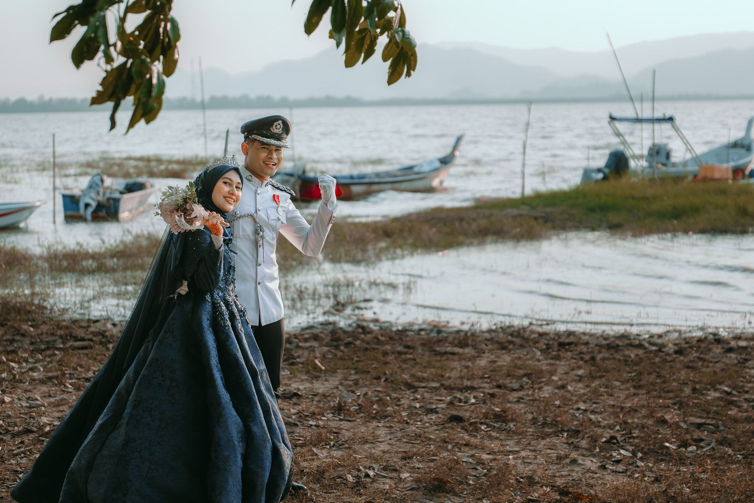 Two figures in formal attire pose near a waterfront with boats and mountains visible in the background.