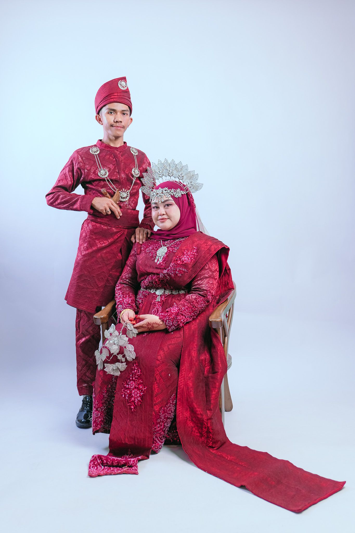 Two people wearing traditional red Malaysian wedding attire pose together against a light background.