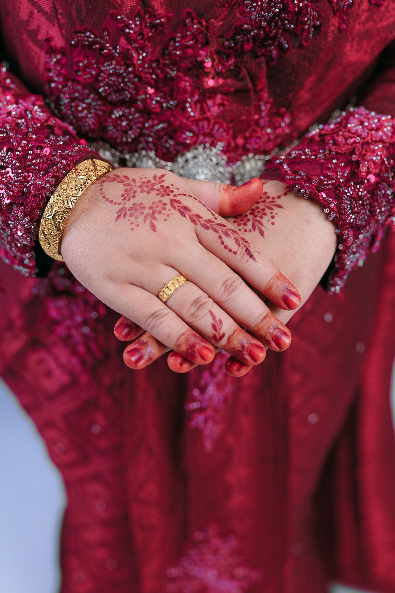 Detailed close-up of hands with henna tattoo and gold jewelry against a deep red embellished fabric.