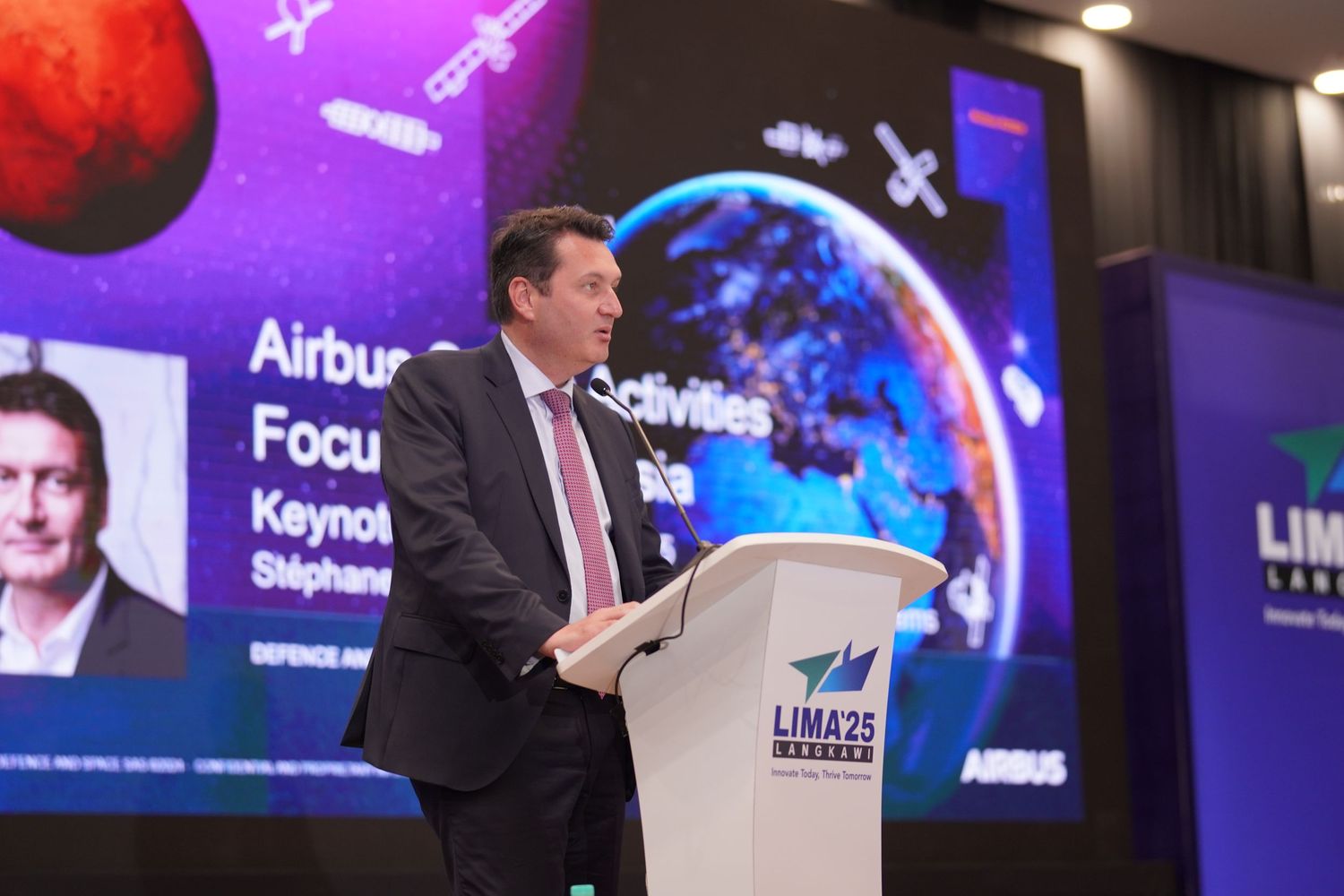 Speaker presents at podium with space-themed graphics displayed on screen behind.