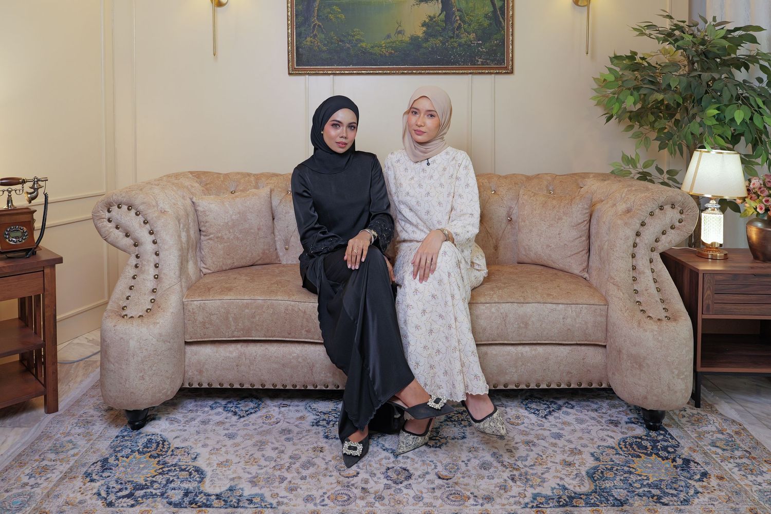 Two people sit together on a beige couch in a traditional living room with a patterned area rug and table lamp.