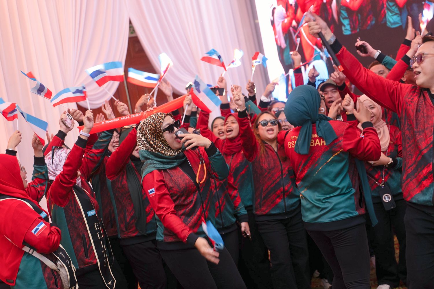 A large group of women in matching red uniforms wave flags and celebrate enthusiastically at an outdoor event.