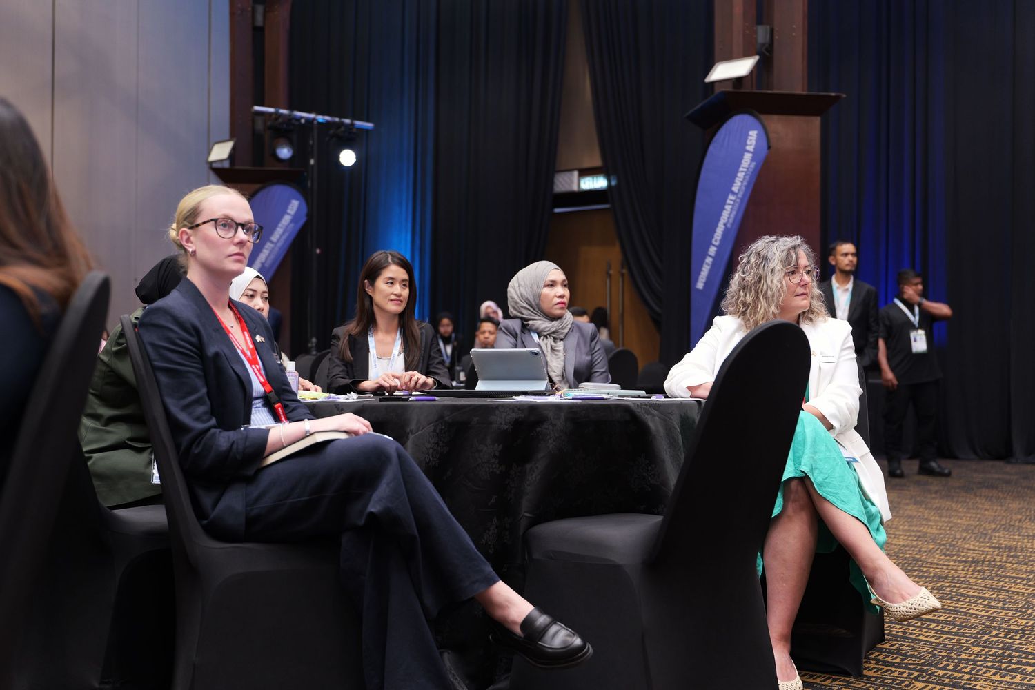 Professional conference setting with blue stage lighting and attendees seated at round tables.