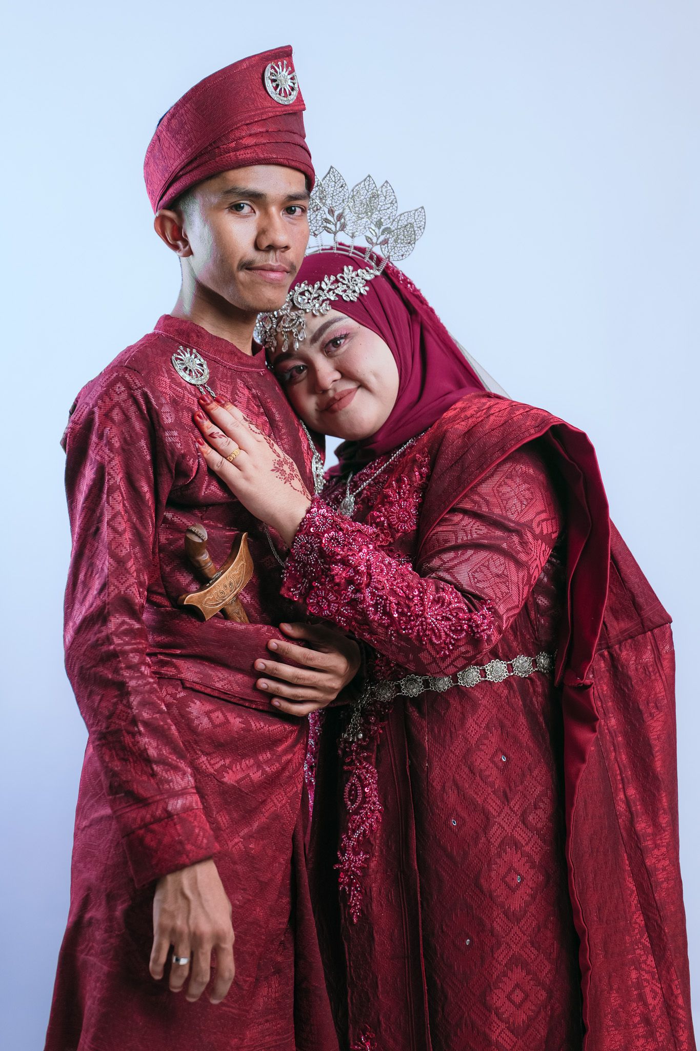 A couple embraces lovingly in matching deep red traditional attire with decorative embellishments against a light backdrop.