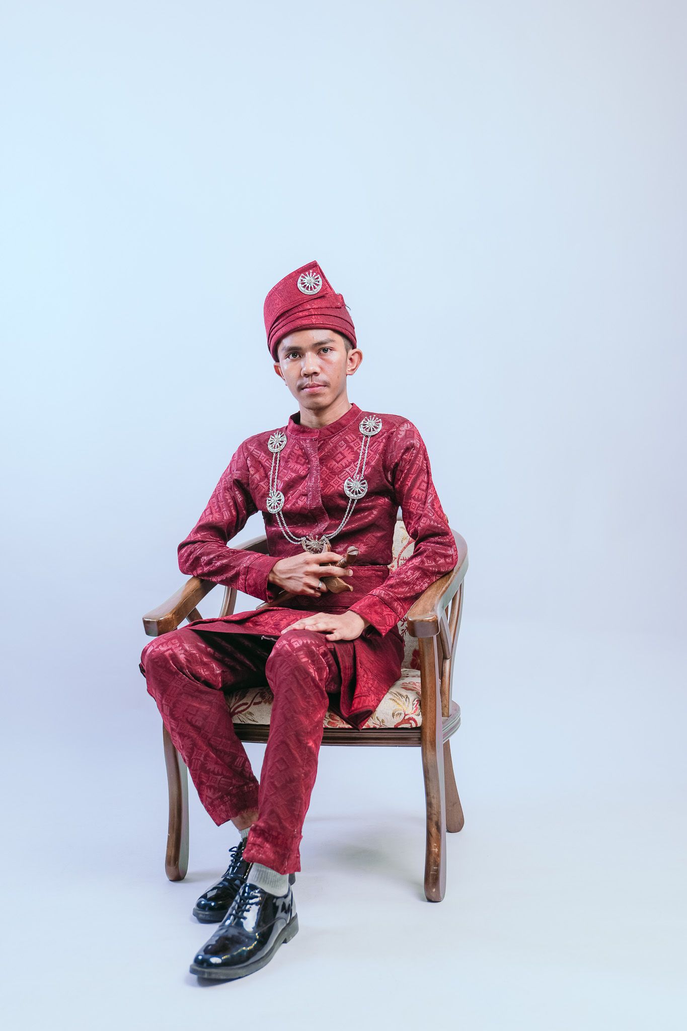 Man in traditional red Malay formal attire sitting on wooden chair holding ceremonial dagger.