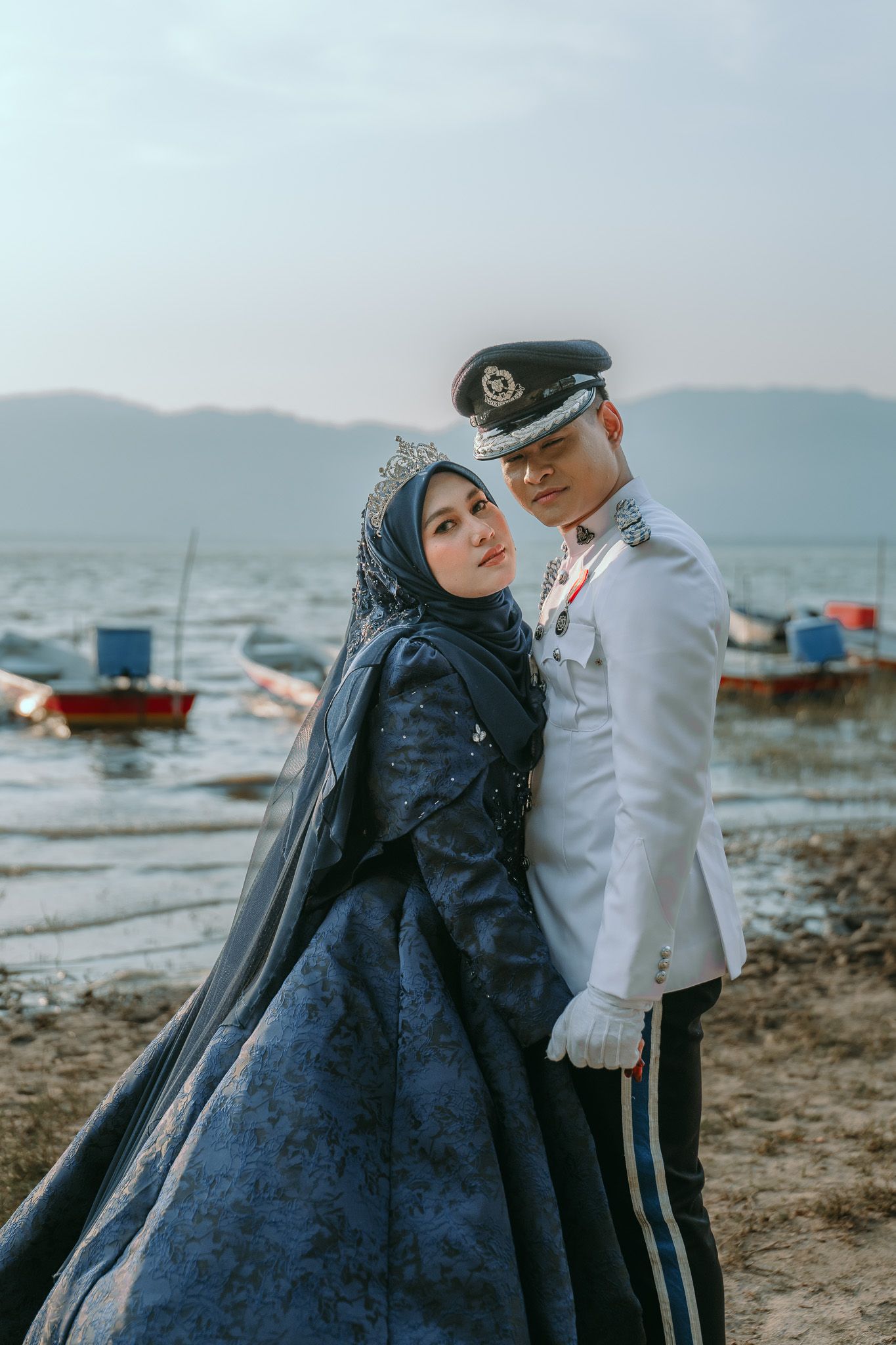 A couple embraces by a seaside harbor with boats in the background, wearing formal military dress and flowing navy gown.