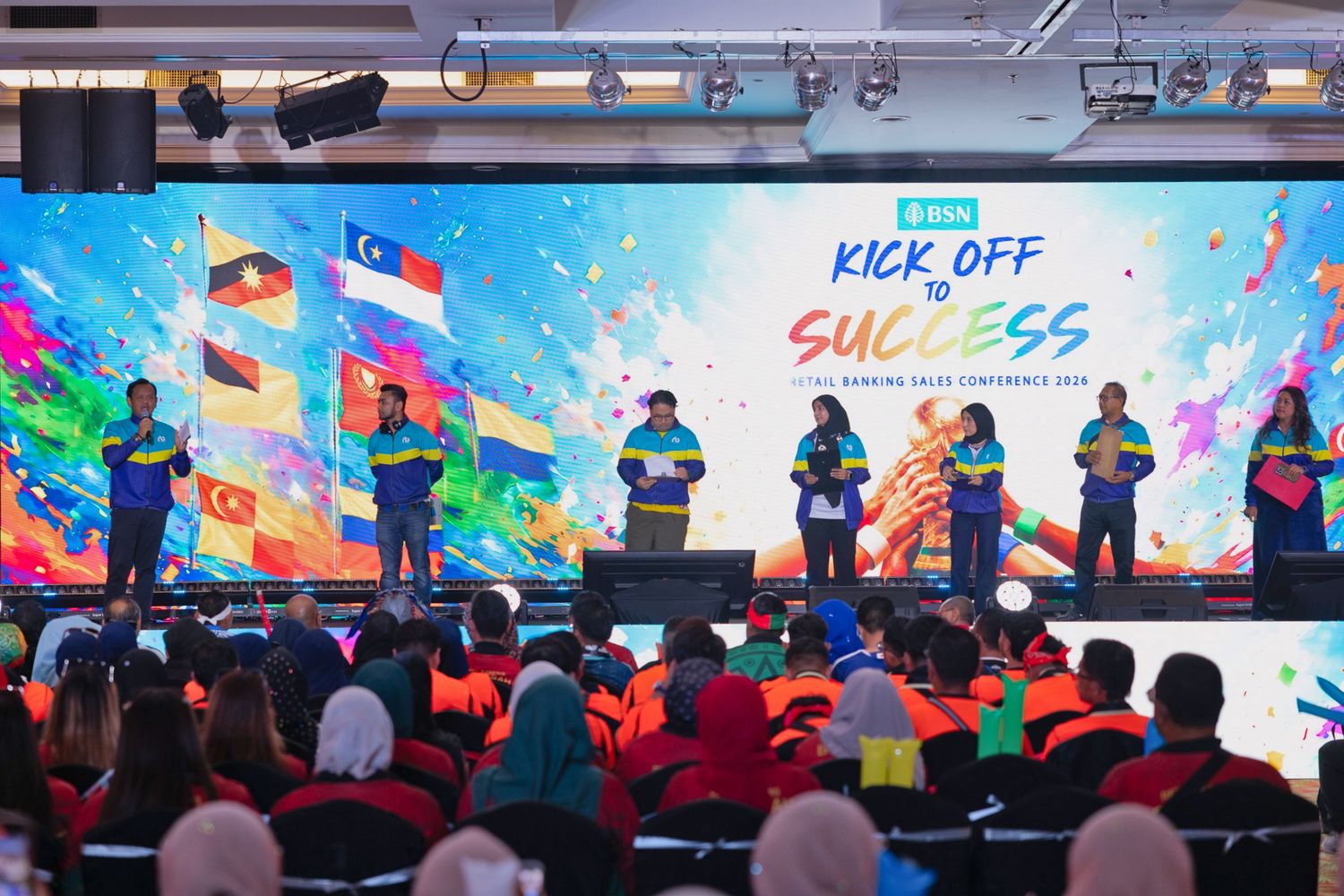 Corporate conference stage displays Kick Off to Success banner with speakers and colorful flags in a packed auditorium.